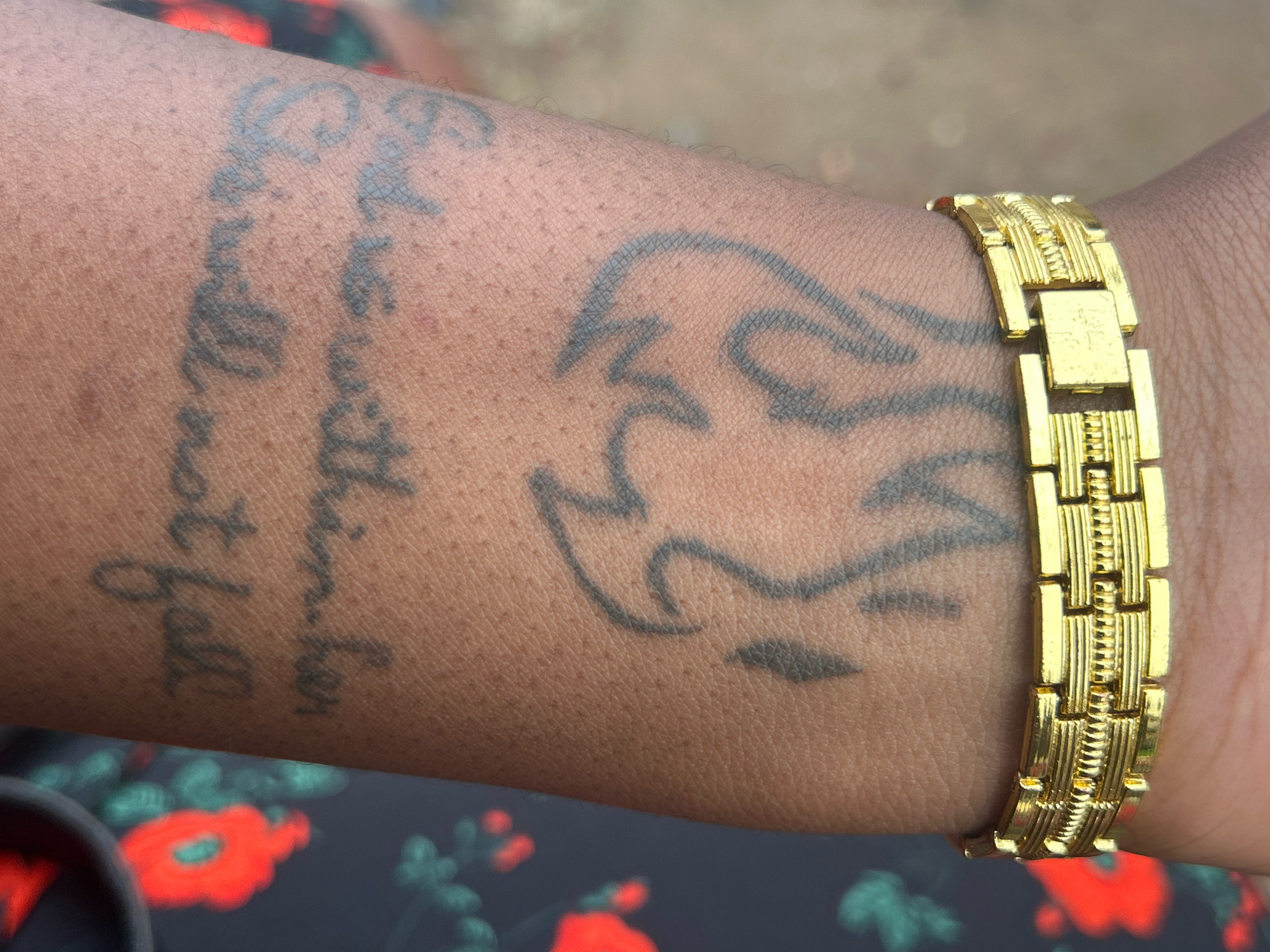 The image shows a close-up of a person's forearm in Ikotun, Nigeria. A gold bracelet is worn on the wrist. The forearm has two tattoos: one with script that appears to say "God will not fall," and another abstract design resembling a flame or wings. The skin texture and pores are visible. The background is blurred but shows a dark fabric with a floral pattern of red and green. The lighting suggests an outdoor setting.