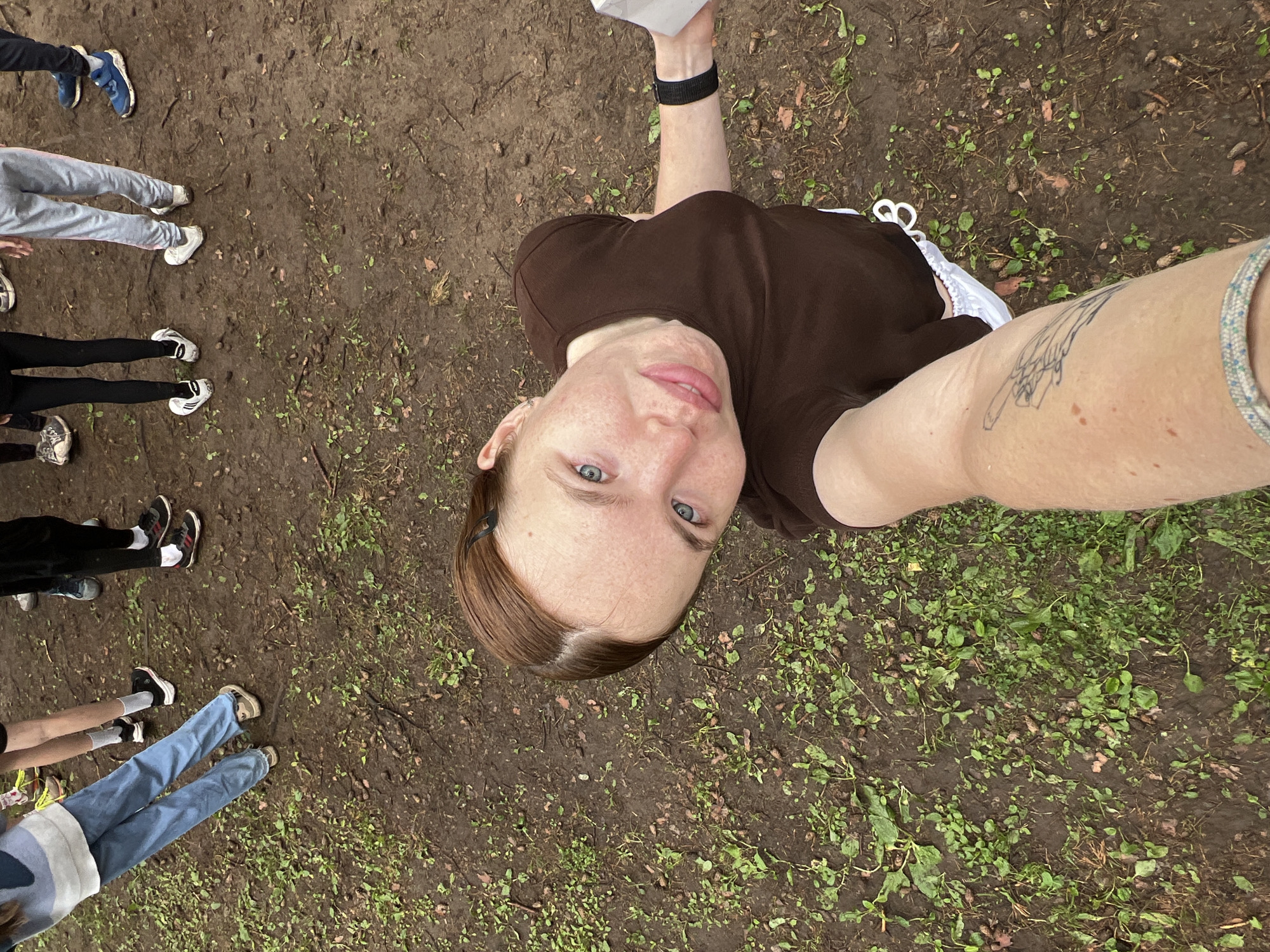 The image shows a young woman with auburn hair and freckles taking a selfie from a high angle, looking up at the camera. She is wearing a brown t-shirt and white shoes. Her left arm, tattooed with a wing design, is extended towards the camera. In the background, several other individuals are standing on a muddy ground with sparse green vegetation. Their legs and feet are visible, suggesting they are wearing casual clothing and sneakers. The environment appears to be an outdoor area, possibly a park or a trail, with damp, earthy soil and scattered leaves. No specific landmarks or recognizable features of Chaikovsky, Russia, are identifiable in this image. The scene is otherwise unremarkable, depicting a casual outdoor gathering.
