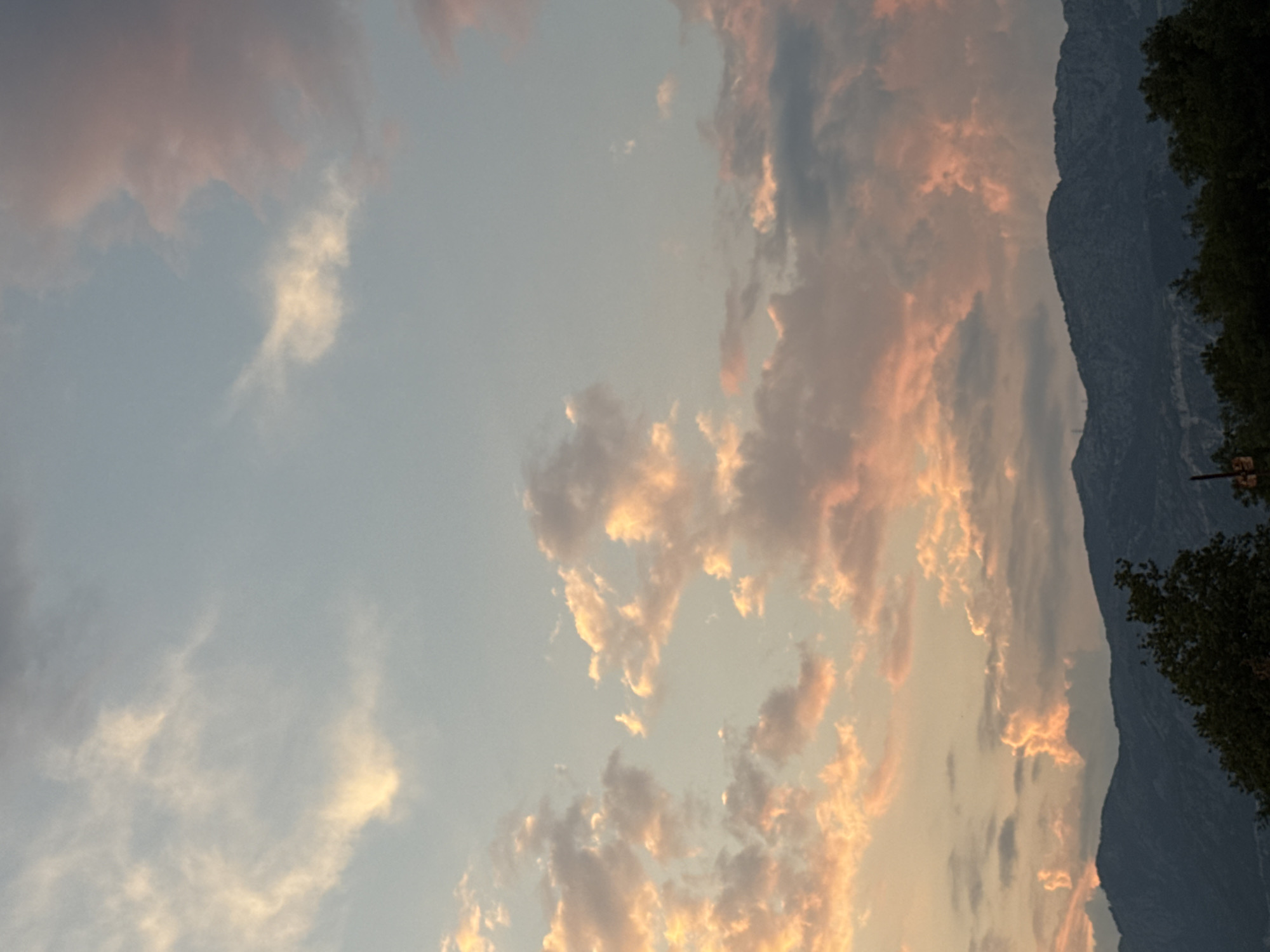 This image captures a serene evening sky over a mountainous landscape, likely in Kamenovo, Montenegro. The primary subjects are the clouds, painted in hues of soft pink, orange, and gray, reflecting the warm glow of the setting sun. These clouds are scattered across a pale blue sky, with some appearing wispy and translucent while others are more dense and textured.
To the right of the frame, a dark, rugged mountain range rises dramatically, its slopes covered in the shadows of dusk. The silhouette of the mountain is sharp against the lighter sky. In the foreground, on the far right, the dark green foliage of trees can be seen, suggesting a natural setting. The overall scene evokes a sense of tranquility and the beauty of nature during twilight.
The time of day is clearly sunset or twilight, as indicated by the warm, diffused lighting and the colors of the clouds. The weather appears calm, with no strong winds suggested by the cloud formations. The location cues, such as the mountain range and the hint of vegetation, are consistent with a coastal or mountainous region. No people or specific objects are clearly discernible in the image, focusing the viewer's attention on the natural elements. No text is visible.