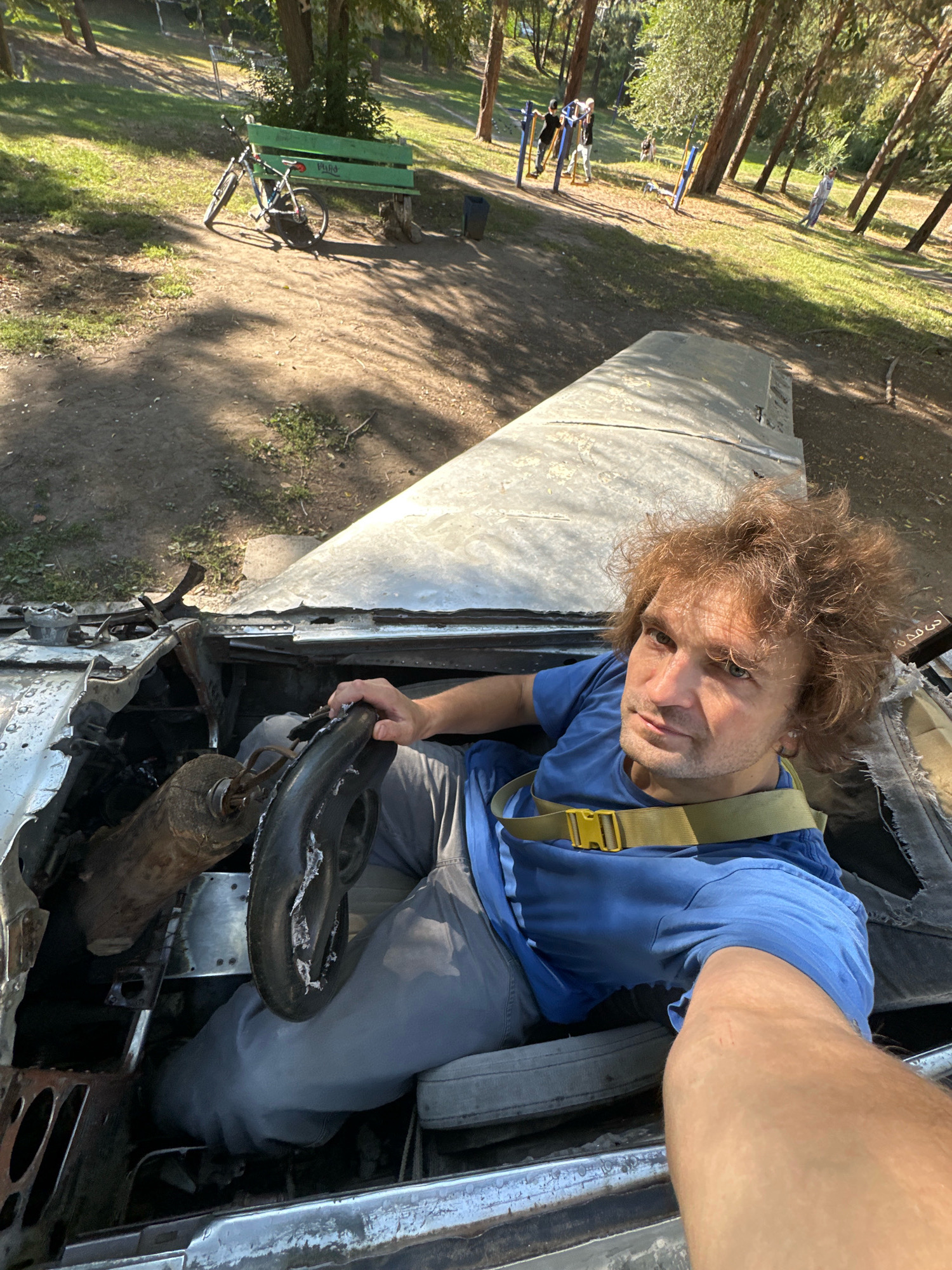 A man is sitting in the cockpit of what appears to be a wrecked vehicle, possibly an airplane, in a park. He is wearing a blue t-shirt, grey pants, and a yellow strap across his chest. His left hand is on the steering wheel. The cockpit is damaged and appears weathered. In the background, a park setting is visible with trees, a green bench, a bicycle, and other people in the distance on what seems to be an outdoor gym. The location is identified as Biysk, Russia.