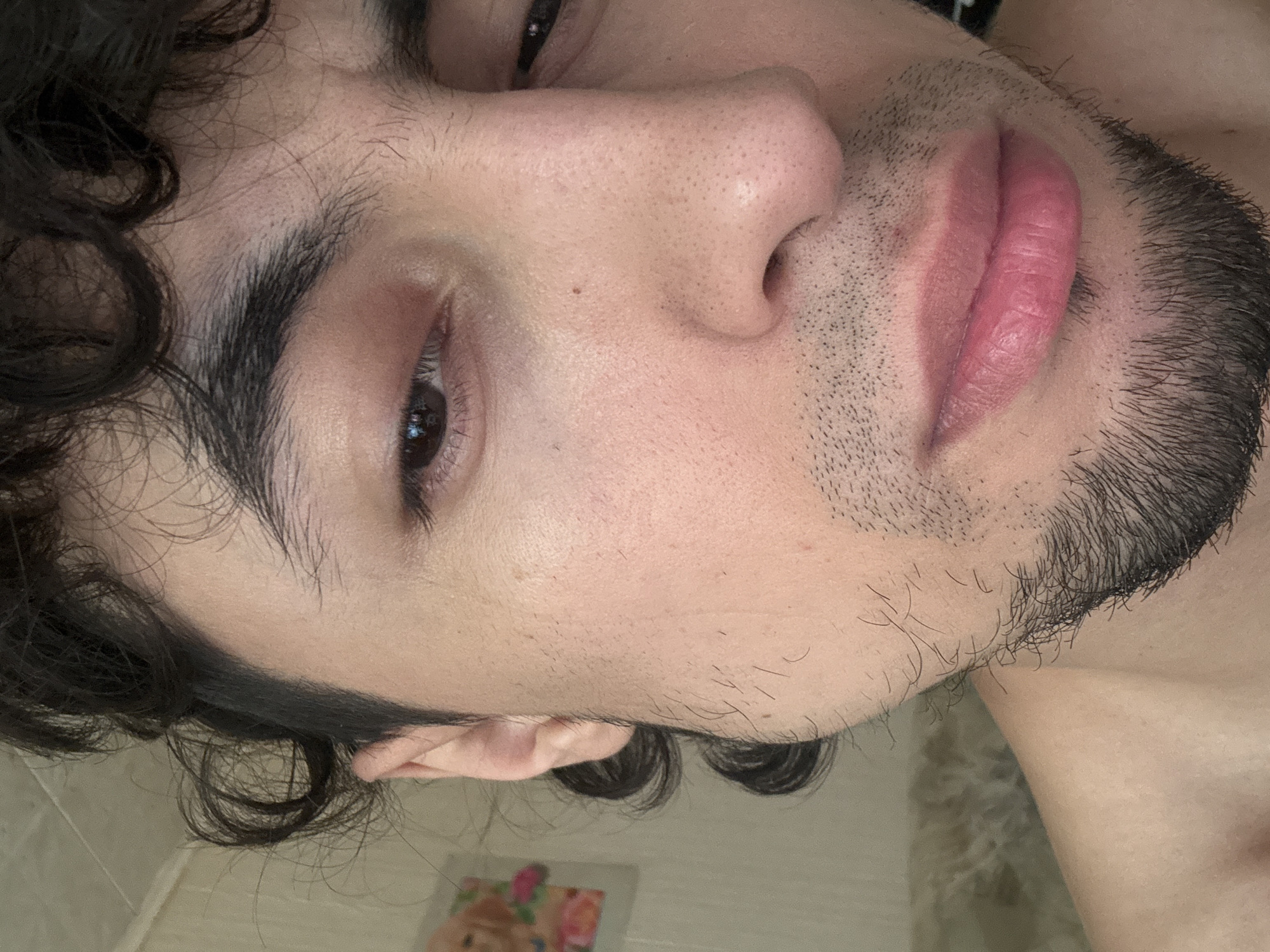 The image is a close-up, low-angle shot of a person's face, specifically focusing on their eye, nose, and lips, with curly dark hair framing the left side of the face. The person appears to have light skin and dark eyebrows and eyelashes. There is a hint of stubble on their chin and jawline. The lighting suggests it could be daytime, possibly indoors given the texture of the background surface which resembles a tiled wall. The person's lips are slightly parted and appear to be a natural pink color. The overall mood is intimate and personal, with a focus on the facial features. There is no visible text in the image.