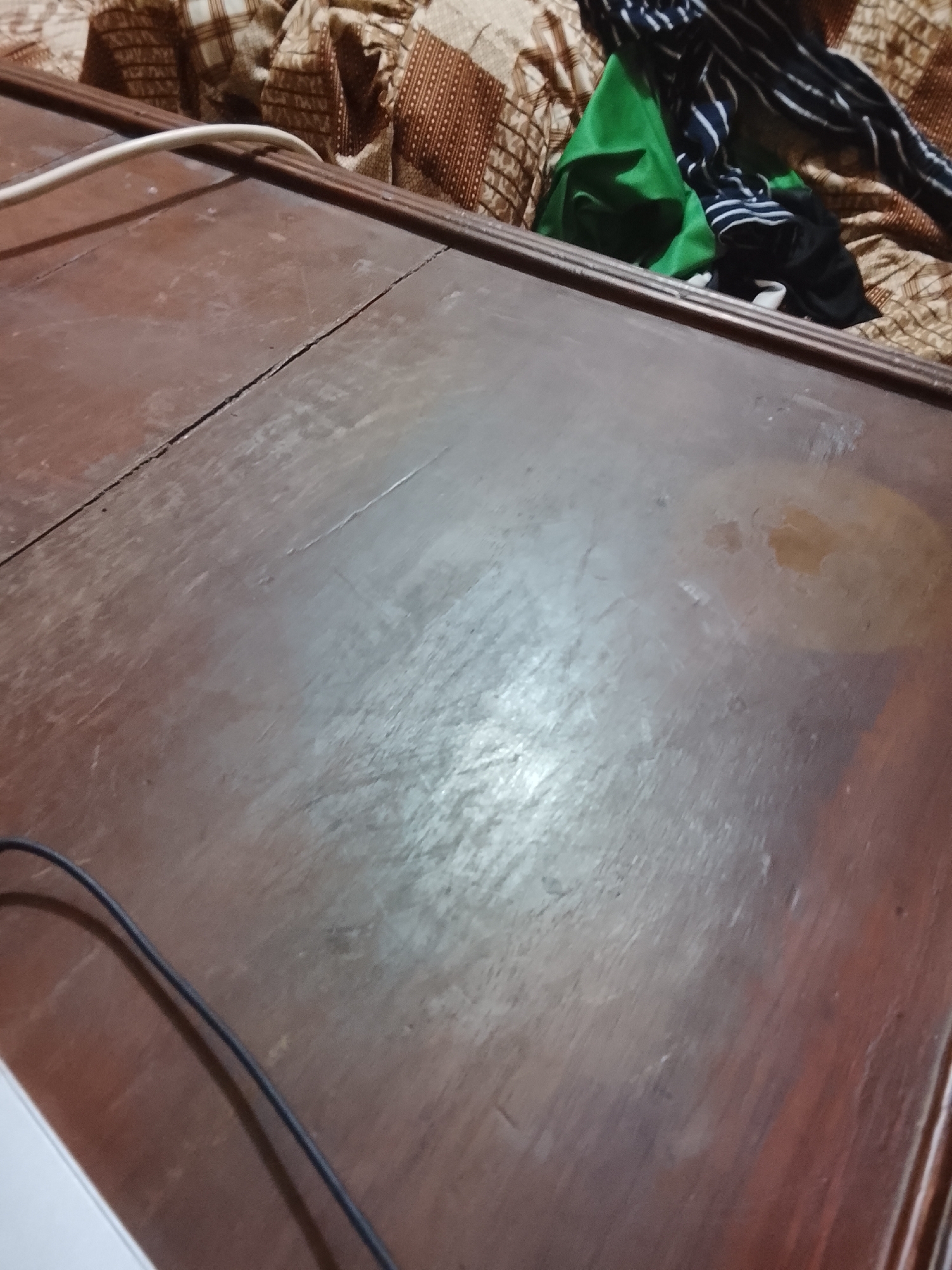 A medium-shot depicts a dark wooden table with several cables running across it. An assortment of clothing items, including green, black, and striped fabrics, are piled on top of patterned fabric in the background. There is no evidence to suggest the city of Multan, Pakistan.