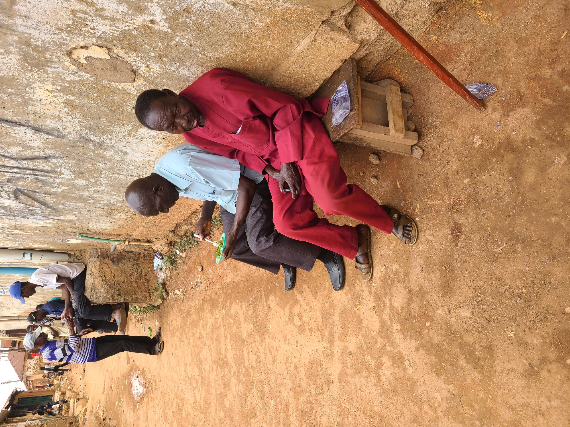 Two men are sitting on a raised concrete structure adjacent to a wall. One man wears a red suit and sandals and leans against the wall. The second man wears a light blue shirt and dark pants, and he holds a small green bowl and a utensil in his hands. The ground is dirt. In the background, other people are sitting and standing along a street. The image suggests the scene is outdoors in an urban environment. Based on context, the location is Jos, Nigeria.