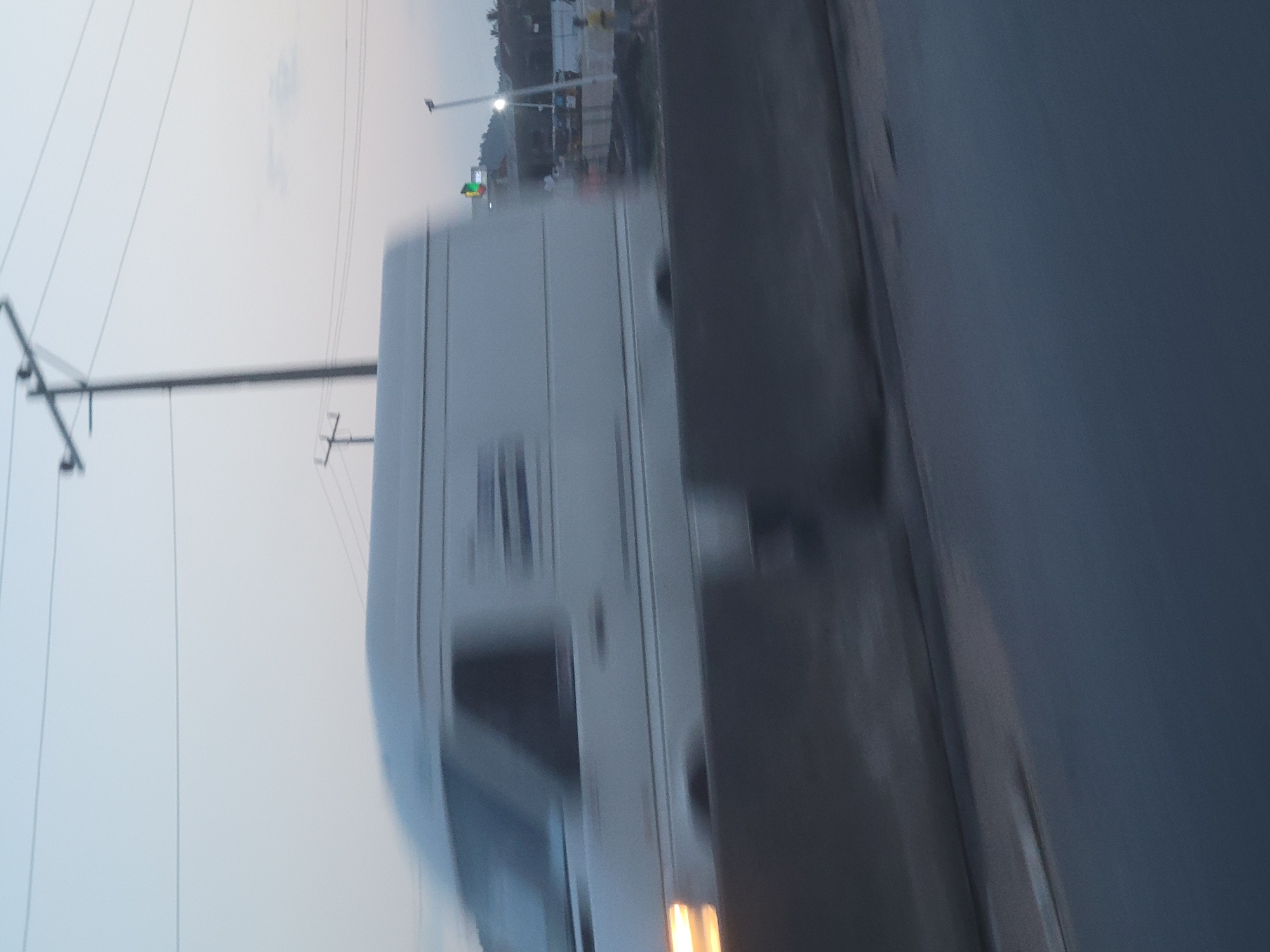 The media file depicts a moving vehicle, likely a bus or a large truck, on a road. The camera is angled, giving a sense of motion and speed. The sky is overcast, suggesting it could be dawn or dusk, or a generally cloudy day. In the background, there are signs of human activity, with what appears to be a traffic light and some structures that could be buildings or overpasses. The overall scene is urban or suburban.
The main subject is the side of the vehicle, which is a light color and appears to have several windows. The motion blur is significant, indicating the photograph was taken from another moving vehicle or that the vehicle itself is moving at a high speed. There are power lines visible in the sky, a common sight in developed areas. The road surface is dark and appears to be asphalt.
The lighting is subdued, consistent with the overcast sky. There are no clearly discernible people in the image, but the presence of the vehicle and traffic signals implies a populated area. The environment appears to be a highway or a major road. The time of day is difficult to determine precisely due to the lighting, but it's likely not the middle of the day. No text is visible in the image.