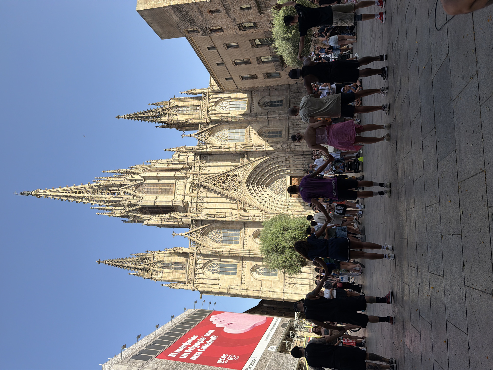 The image shows a group of people holding hands in a circle in front of the Barcelona Cathedral, located in the Gothic Quarter of Barcelona, Spain. The cathedral is a large Gothic structure with intricate spires and architectural details, bathed in sunlight. The people are diverse in age and gender, with some dressed casually in shorts and t-shirts, while others are in more formal attire. Some individuals in the foreground are wearing athletic wear. The ground is paved with large, square stone tiles. A large red billboard with text and an image of a pink foot is visible on the left side of the frame, adjacent to a building under construction. In the background, more people are gathered, observing the scene, and there are trees and other buildings. The sky is clear and blue. The overall atmosphere appears to be one of community and celebration.