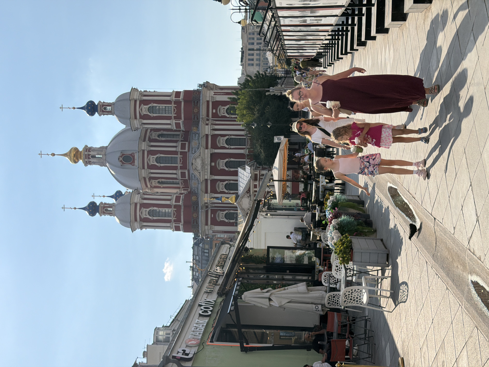 The image depicts a street scene in Moscow, Russia, likely on a sunny day, with the facade of a large, red-and-white church with multiple domes and crosses prominently visible in the background. The foreground features a pedestrian walkway paved with light-colored square tiles. A narrow, U-shaped channel runs along the walkway, containing water that reflects the sky. A black bird is present in the channel.
Several people are walking along the walkway. In the immediate foreground, two young girls are walking side-by-side. The girl on the left is wearing a pink tank top and patterned shorts. The girl on the right is wearing a pink t-shirt and a pink and white skirt. Behind them, a woman in a dark maroon dress is walking, holding a cup. Another woman in a white top and hat is walking behind the girls. Further back, more people are visible walking and seated at outdoor cafes.
To the left of the walkway is a row of buildings with outdoor seating areas. One building has a green facade with an awning and signs in Russian. Several tables and chairs are set up outside. Flower pots with colorful flowers are placed along the edge of the walkway.
To the right of the walkway, a series of display boards or posters are set up on stands, forming a long row. The sky is blue with a few wispy clouds. The overall atmosphere appears to be that of a vibrant, pedestrian-friendly urban area.