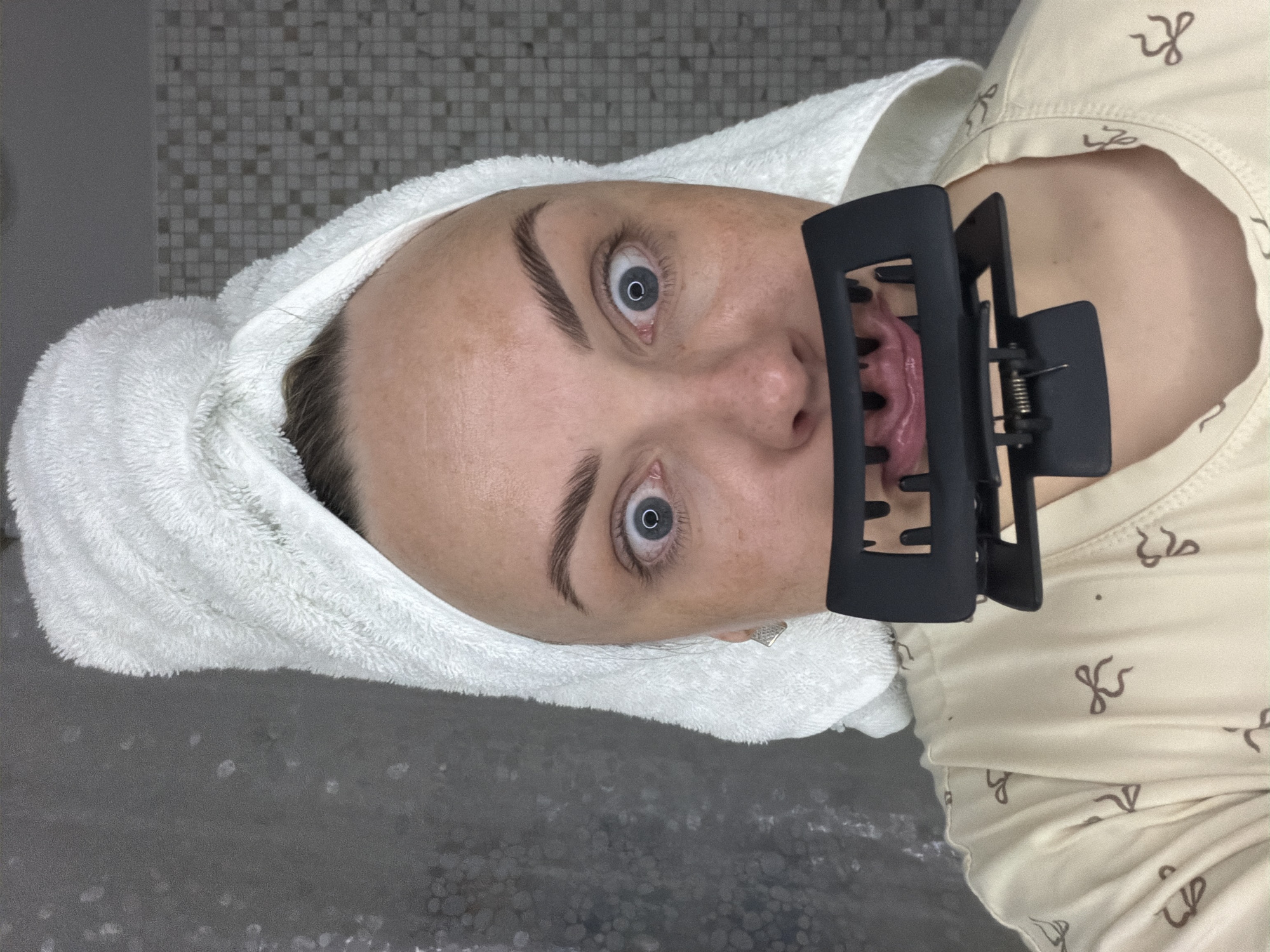 A person with a white towel wrapped around their head is photographed close-up, likely indoors. A black hair clip is placed over their mouth. The person's eyes are wide open. They are wearing a cream-colored shirt with a brown pattern. The background features a tiled wall and a possibly wet surface. The image does not contain any specific details identifying the location as Votkinsk, Russia.