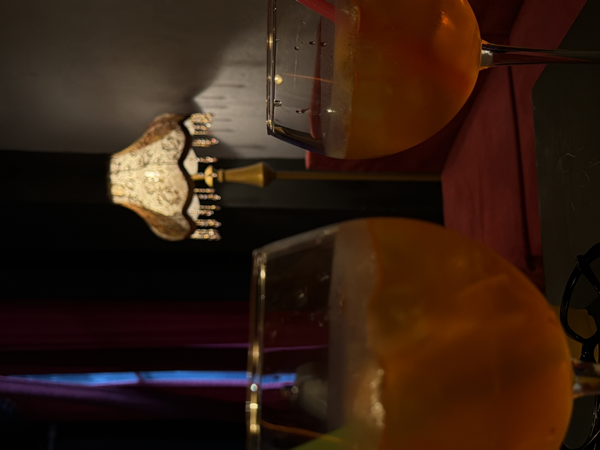 Two glasses of an orange beverage, likely cocktails, are in the foreground, with one glass slightly out of focus. The glasses are filled with ice and appear to have garnishes or straws. In the background, a vintage-style lamp with a patterned shade and beaded fringe casts a warm glow. Behind the lamp, a dark red curtain hangs, and to the left, a glimpse of what appears to be a blue light is visible. The scene suggests an indoor setting, possibly a bar or lounge. There are no people or animals visible in the image. The location is not discernible from the image itself.