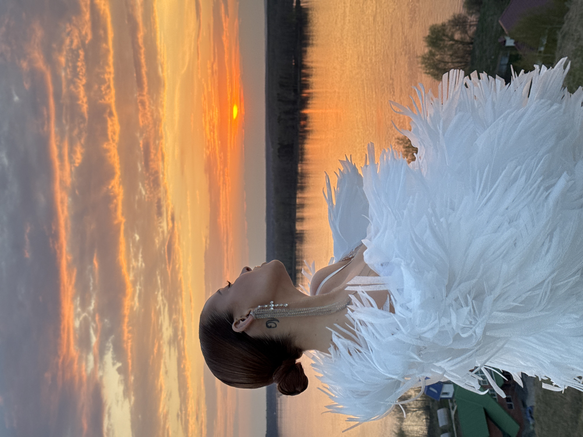 The image depicts a woman in a white feathered garment, possibly a dress or cape, standing outdoors. She is facing away from the camera, with her head tilted slightly upward towards the sky. Her hair is styled in a bun, and she is wearing a long, ornate earring on her left ear. A tattoo is visible on the back of her neck.
The background features a sunset over a body of water, likely a lake or river. The sky is filled with vibrant orange and yellow hues, reflecting on the water's surface. On the far side of the water, there is a strip of land with some trees. Closer to the woman, there are some buildings with green roofs.
The location appears to be Ольховка, Russia, as indicated in the prompt.