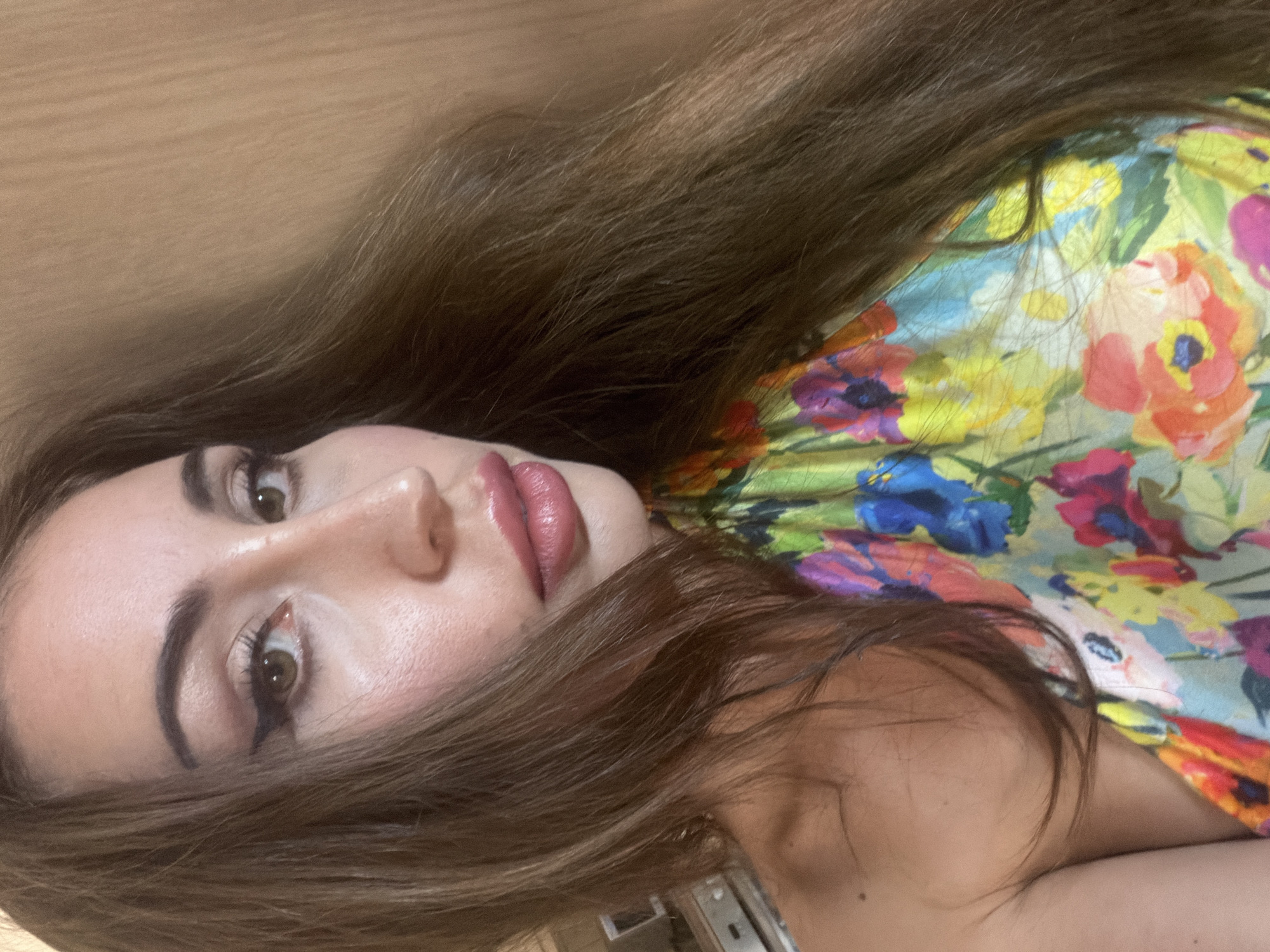This is a close-up, indoor portrait of a woman with long, brown hair. She is wearing a colorful floral top and has on dramatic makeup, including winged eyeliner and lip gloss. The background appears to be a wooden wall. The image does not contain any information about the location being Tersefanou, Cyprus.