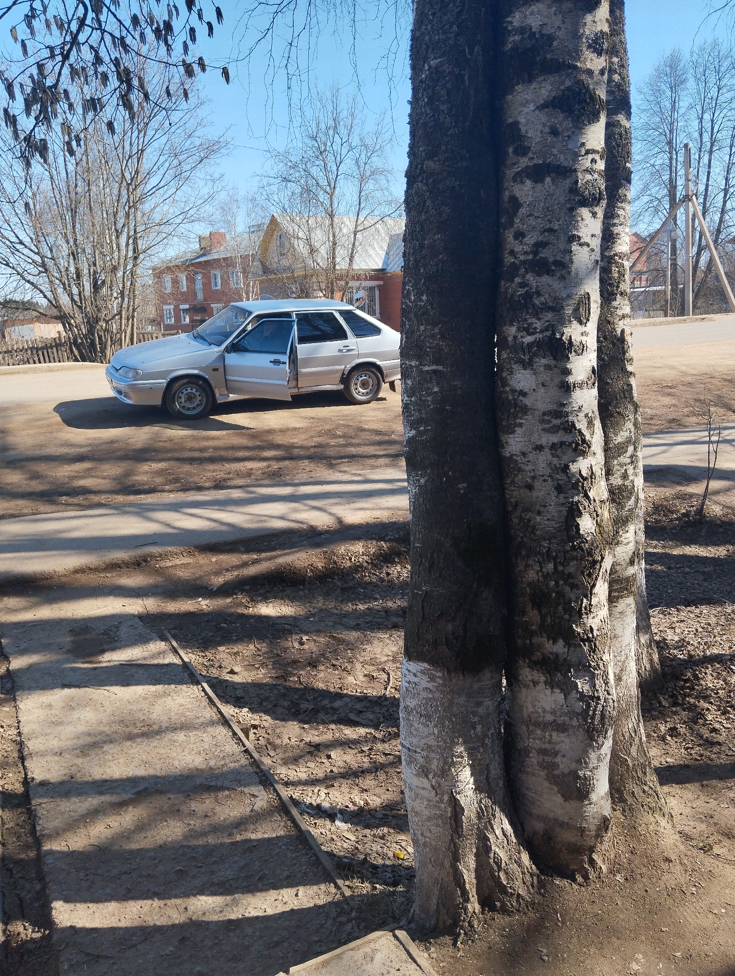 The image shows an outdoor scene in Krasnogorskoye, Russia. A silver Lada sedan with the driver's side door open is parked on a paved road. Behind the car, a two-story brick building and other residential structures are visible. In the foreground, a cluster of three birch trees with partially whitewashed trunks obscures part of the road. The ground consists of a mix of bare earth and a concrete sidewalk section with shadows. The sky is clear and blue.