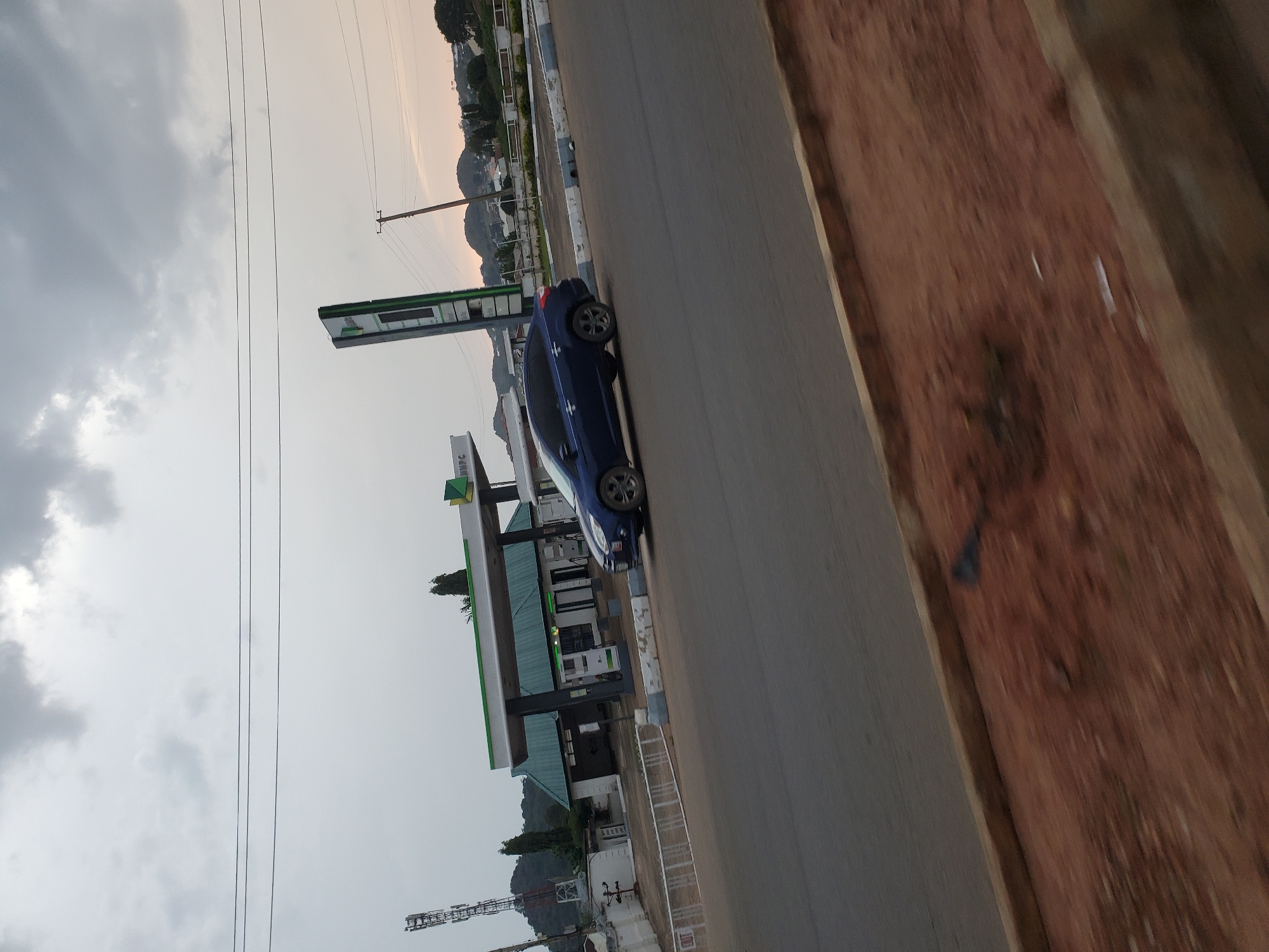 A blue sedan is parked at a gas station in Bukuru, Nigeria. The gas station has a green roof and a tall sign with green accents. Power lines are visible in the background, as are hills and trees. The sky is cloudy.