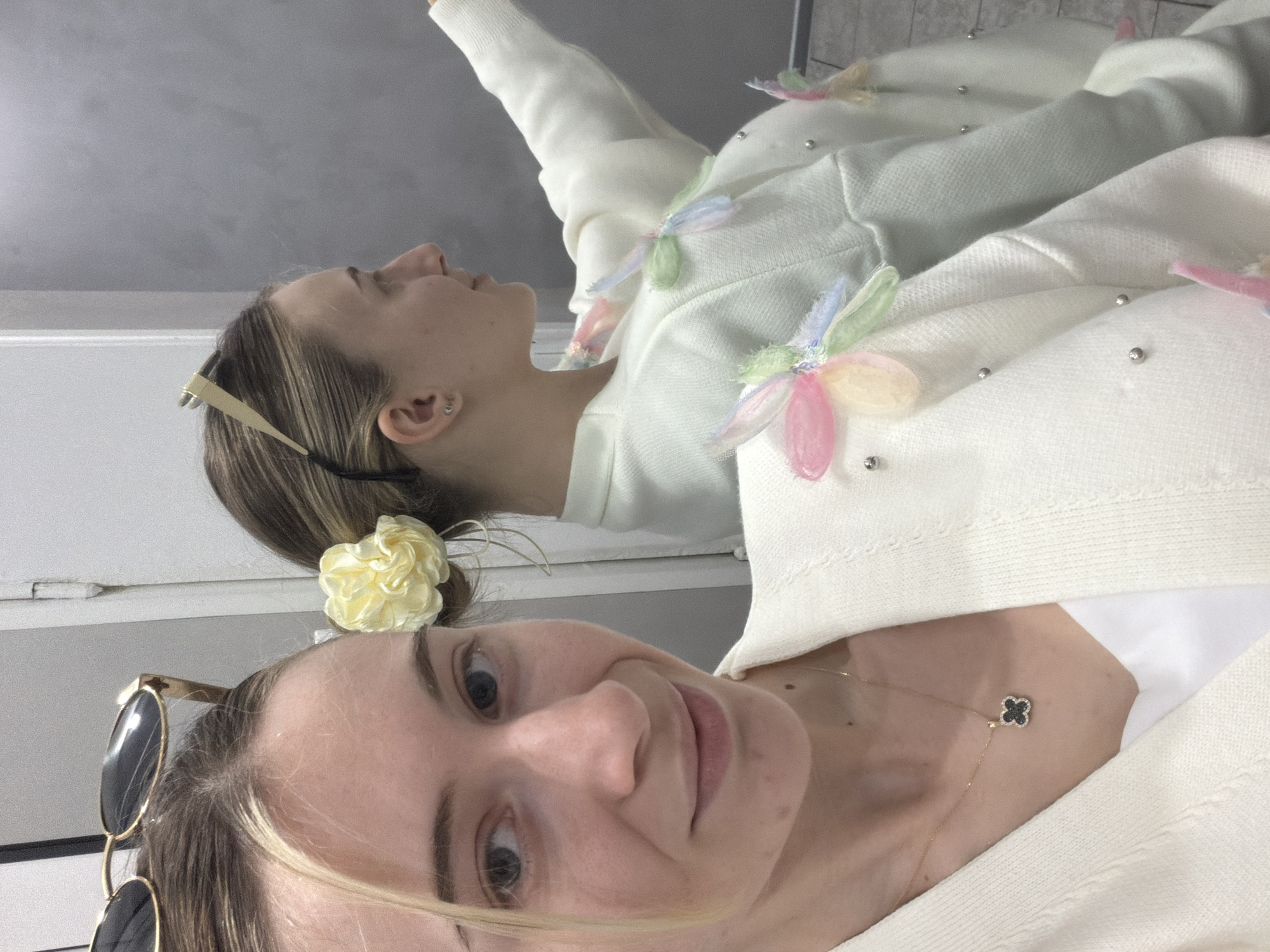 The image shows a woman taking a selfie. She is wearing sunglasses on her head and has a yellow flower in her hair. She also has a gold necklace with a clover pendant. She is wearing a white sweater with flower appliqués. The background appears to be a building interior. There is no information in the image to suggest it was taken in Plovdiv, Bulgaria.