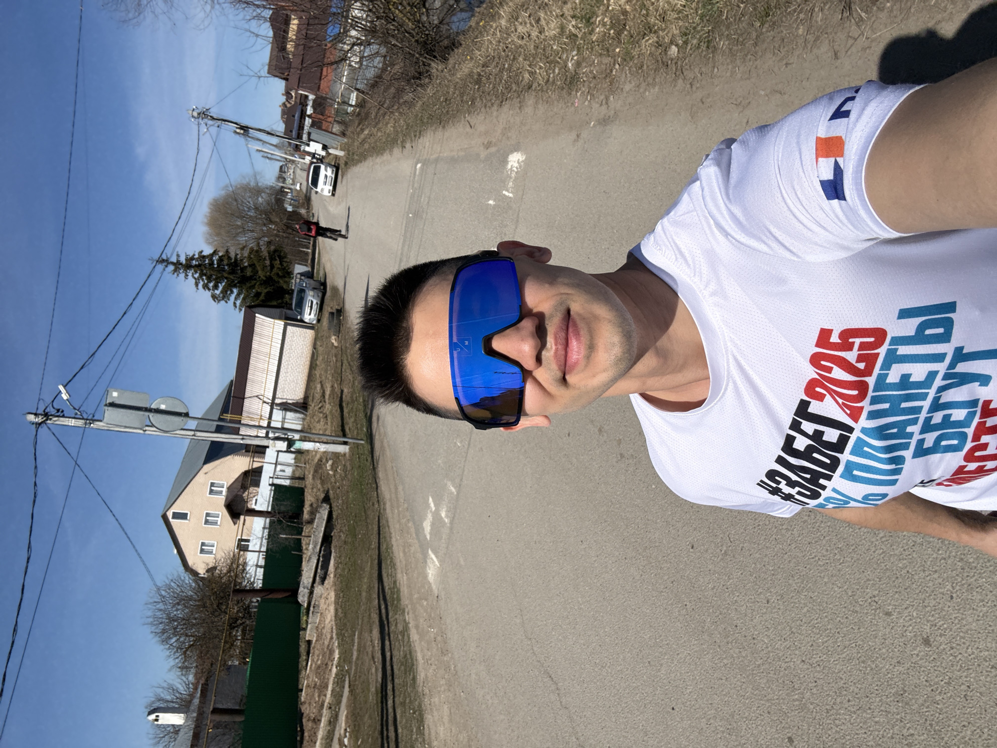 A selfie shows a man with short dark hair and blue mirrored sunglasses standing on a paved road. He wears a white t-shirt with Russian text, including "#ЗАБЕГ2025". The text appears to promote a race event. A house with a green fence is on the left side of the road. Power lines run along the left side. A person walks away down the road. Two cars are parked on the right side. Trees and brush line the road. The blue sky suggests it is daytime. The photo appears to be taken in a residential area. The style of the house suggests it might be a location in Kazan, Russia.
