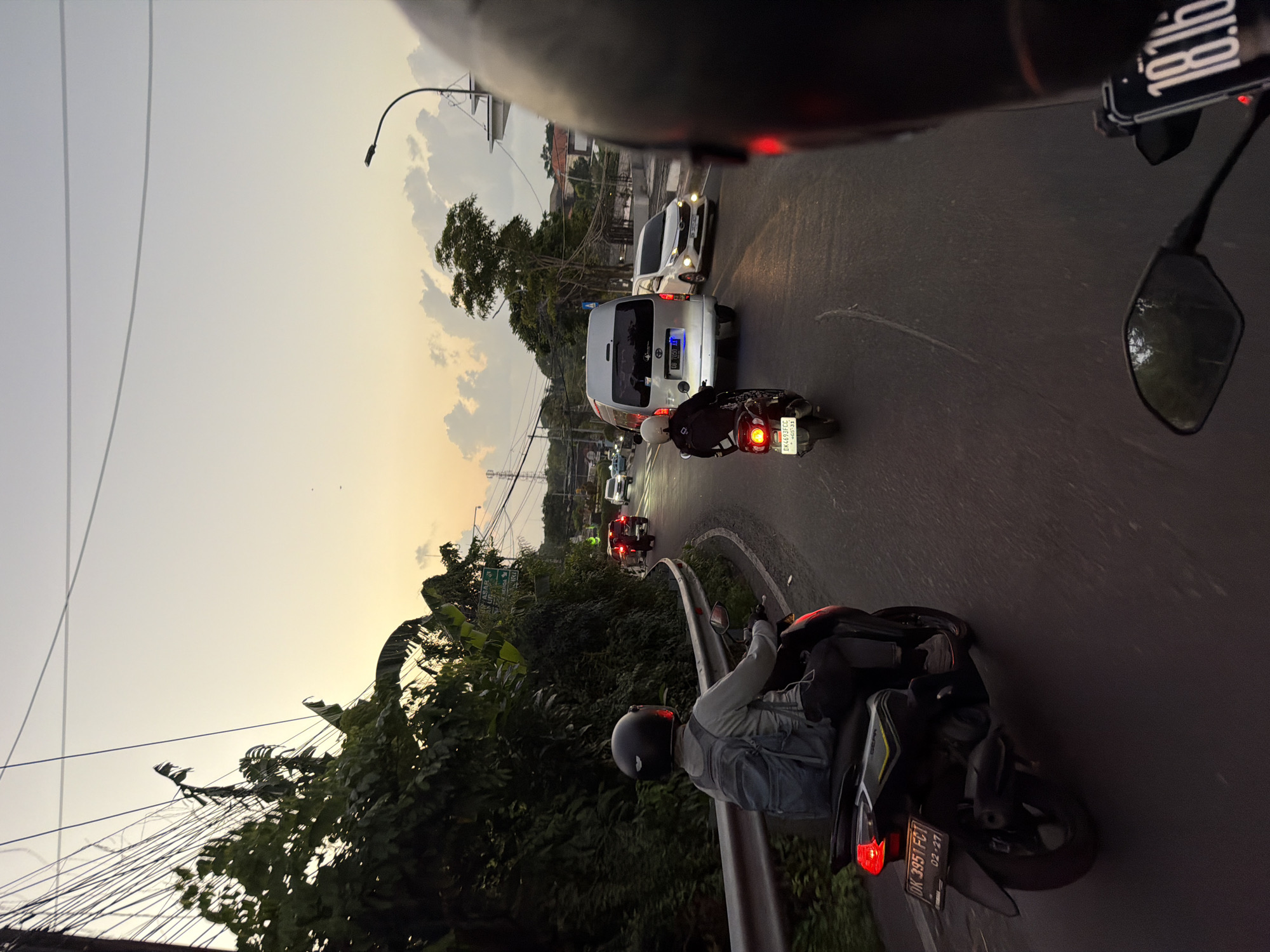 The image depicts a street scene in Kuta Selatan, Indonesia, during the late afternoon or early evening, with a warm, hazy sky. A person wearing a black helmet and a grey jacket with a blue backpack is riding a black scooter on the right side of the road. The scooter has a license plate that reads "DK 3951 FCT 02-27". Another scooter with a rider wearing a white helmet is further ahead, also on the right side of the road, with a license plate reading "DK 4693 FCC 05-33". A silver van with an Indonesian license plate "B 7050 TQ" is in front of the second scooter, and a white car is in the lane next to it. The road is paved and curves to the left. Dense green foliage lines the left side of the road, bordered by a metal guardrail. Utility poles with numerous wires are visible against the sky. In the foreground, the back of another person's helmet is partially visible, suggesting the image is taken from the perspective of a passenger on a third vehicle. The overall atmosphere is one of bustling local traffic in a tropical environment.
