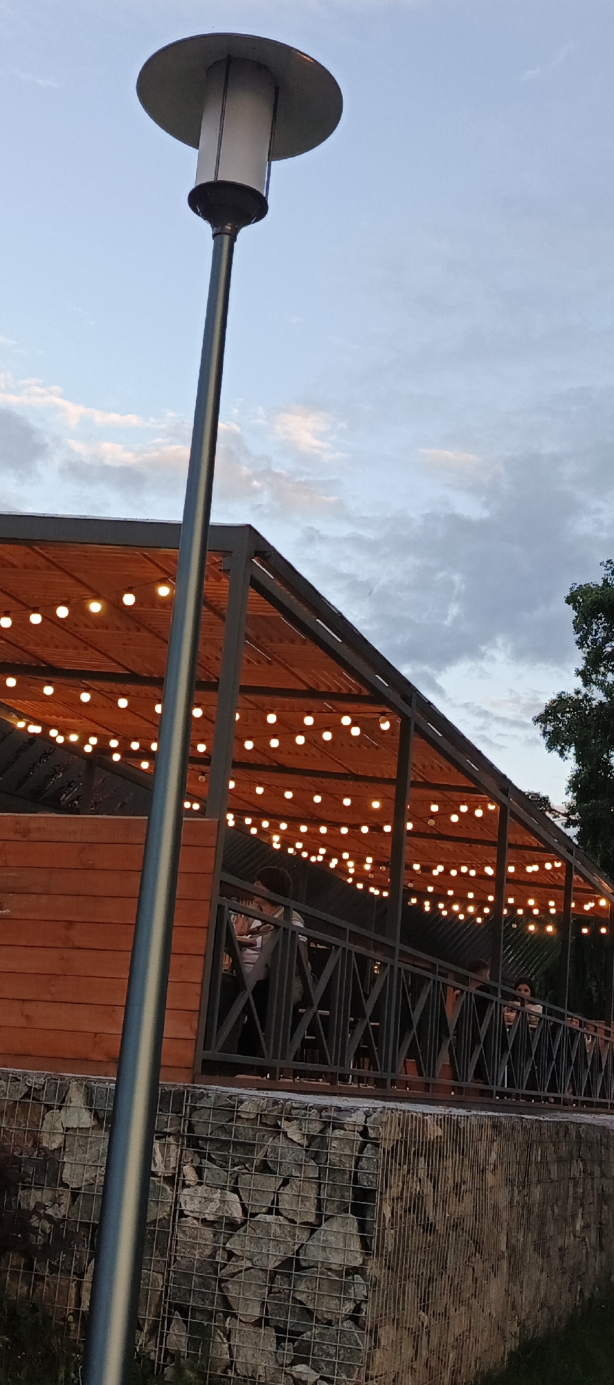 The image depicts an outdoor seating area of a cafe or restaurant in Votkinsk, Russia. In the foreground, a tall, modern lamppost with a silver pole and a cylindrical white light fixture is prominently displayed. Behind the lamppost, a covered patio area is visible, characterized by a wooden ceiling adorned with numerous strings of warm-toned fairy lights. The patio is supported by dark metal beams, and a wooden wall forms the back of the structure. A metal railing with a diamond pattern encloses the patio, and several people are seated at tables within the covered area, some appearing to be dining or socializing. The ground level below the patio is constructed with gabion walls, filled with stones. The sky above is partly cloudy with a blend of blue and grey tones, suggesting it is either early morning or late evening. The overall scene conveys a relaxed, inviting atmosphere, typical of an outdoor dining establishment.