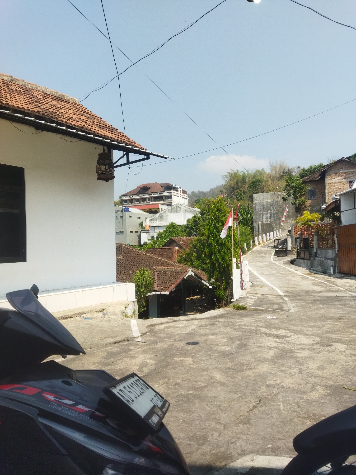A sunlit scene in Wonogiri, Indonesia, features a winding asphalt road ascending a gentle slope. The road is bordered by a concrete barrier on the left and a residential area with terracotta-tiled roofs on the right. Several red and white Indonesian flags are visible along the road. In the foreground, a dark motorcycle is parked, its license plate clearly visible. The background reveals a collection of buildings, including a multi-story structure with a reddish-brown roof, and lush greenery adorning the hillsides. The sky is clear and blue, indicating fair weather.