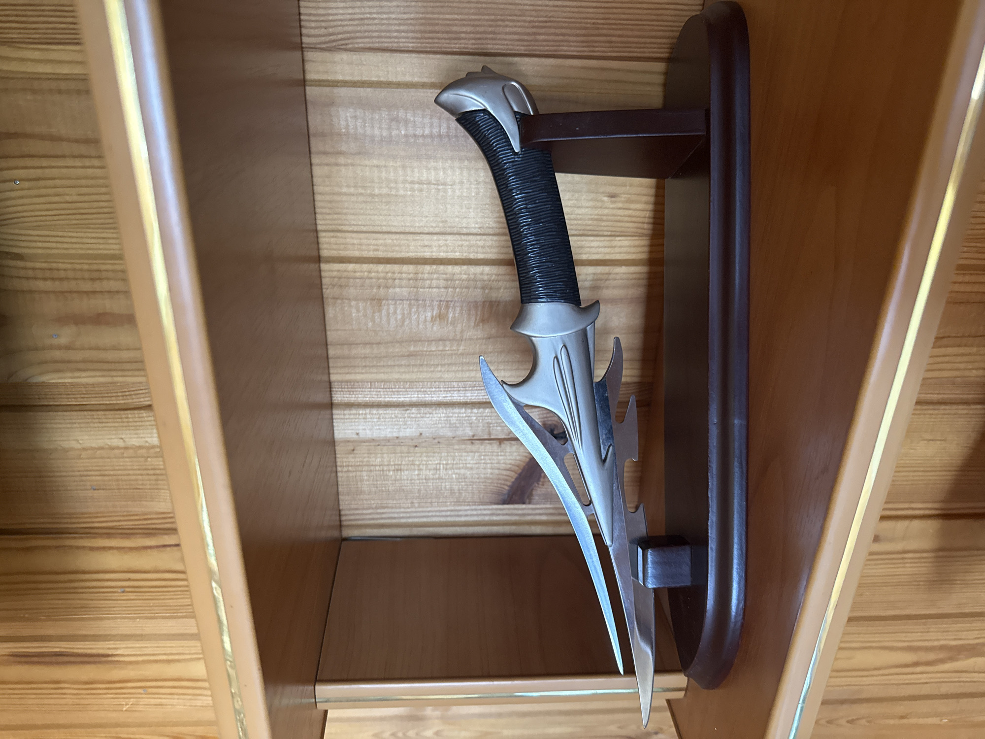 A fantasy-style dagger with a silver, ornate blade and a black, textured handle rests on a dark brown wooden display stand. The stand is on a wooden shelf, which is part of a wooden-paneled wall. The dagger is positioned horizontally, with the blade pointing to the left. The background consists of vertical wooden planks with a light brown tone.