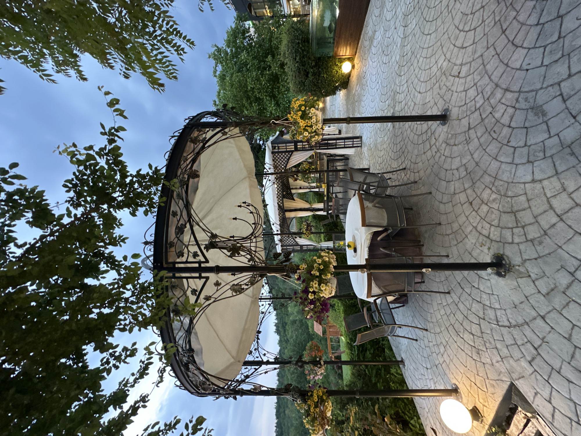 The image depicts an outdoor dining area, likely a restaurant patio, in Veliko Tarnovo, Bulgaria. The scene is set on a paved surface with a circular pattern, possibly cobblestone or stamped concrete. Multiple tables with white tablecloths are visible, surrounded by metal chairs. Each table is shaded by a wrought-iron gazebo-like structure draped with a cream-colored fabric canopy. Hanging flower baskets adorn the structures, adding to the ambiance. Lighting fixtures are situated around the perimeter. The background includes greenery, trees, and potentially parts of other buildings in the city. The image was taken during dusk or twilight.