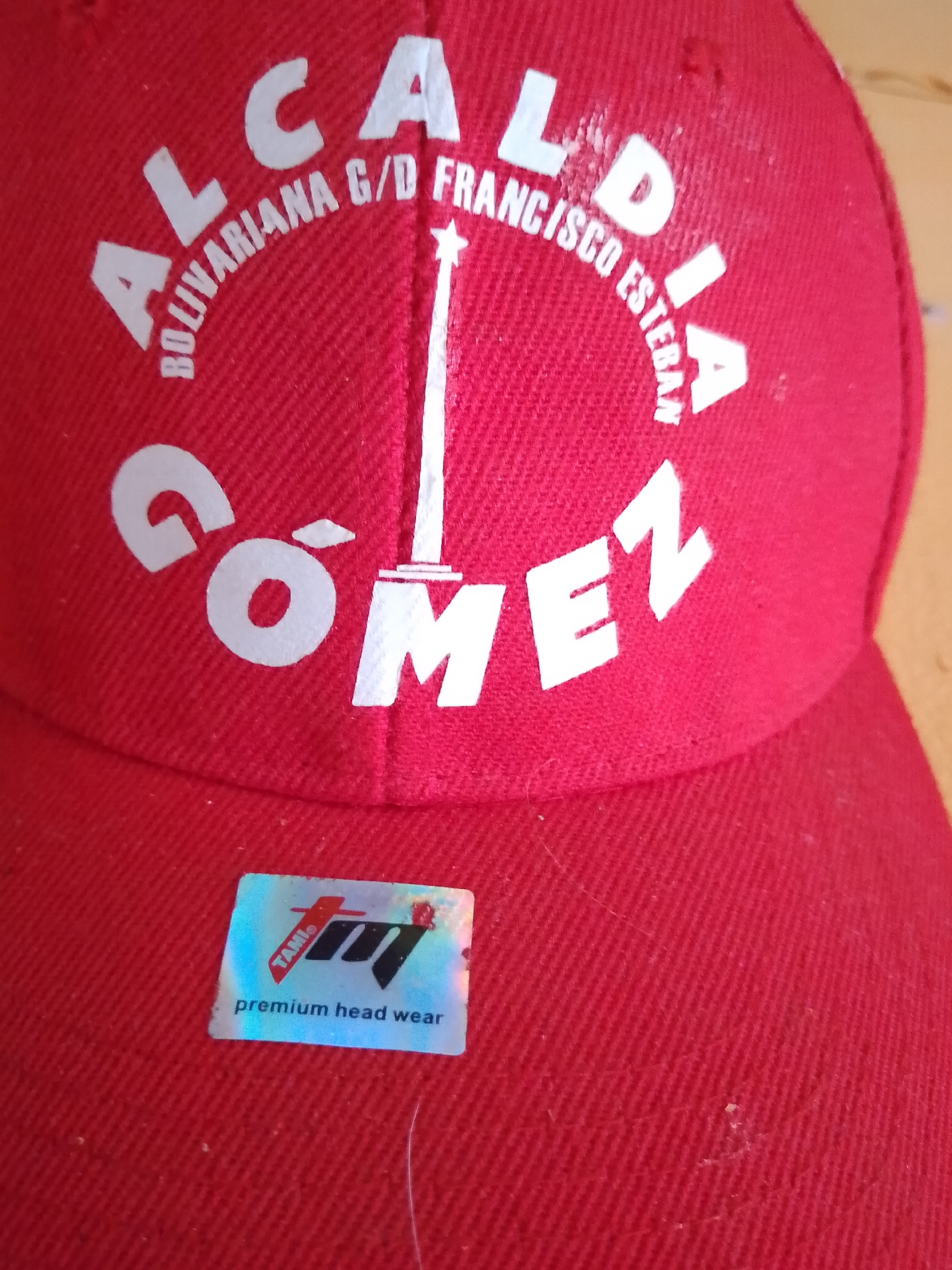 The image shows a close-up of a red baseball cap with white lettering. The lettering on the cap says "ALCALDIA BOLIVARIANA G/D FRANCISCO ESTEBAN GOMEZ". Below this, there is a holographic sticker that says "TAMI. premium head wear". The cap appears to be a souvenir from Altagracia, Venezuela, a city known for its historical significance and association with Francisco Esteban Gómez.
