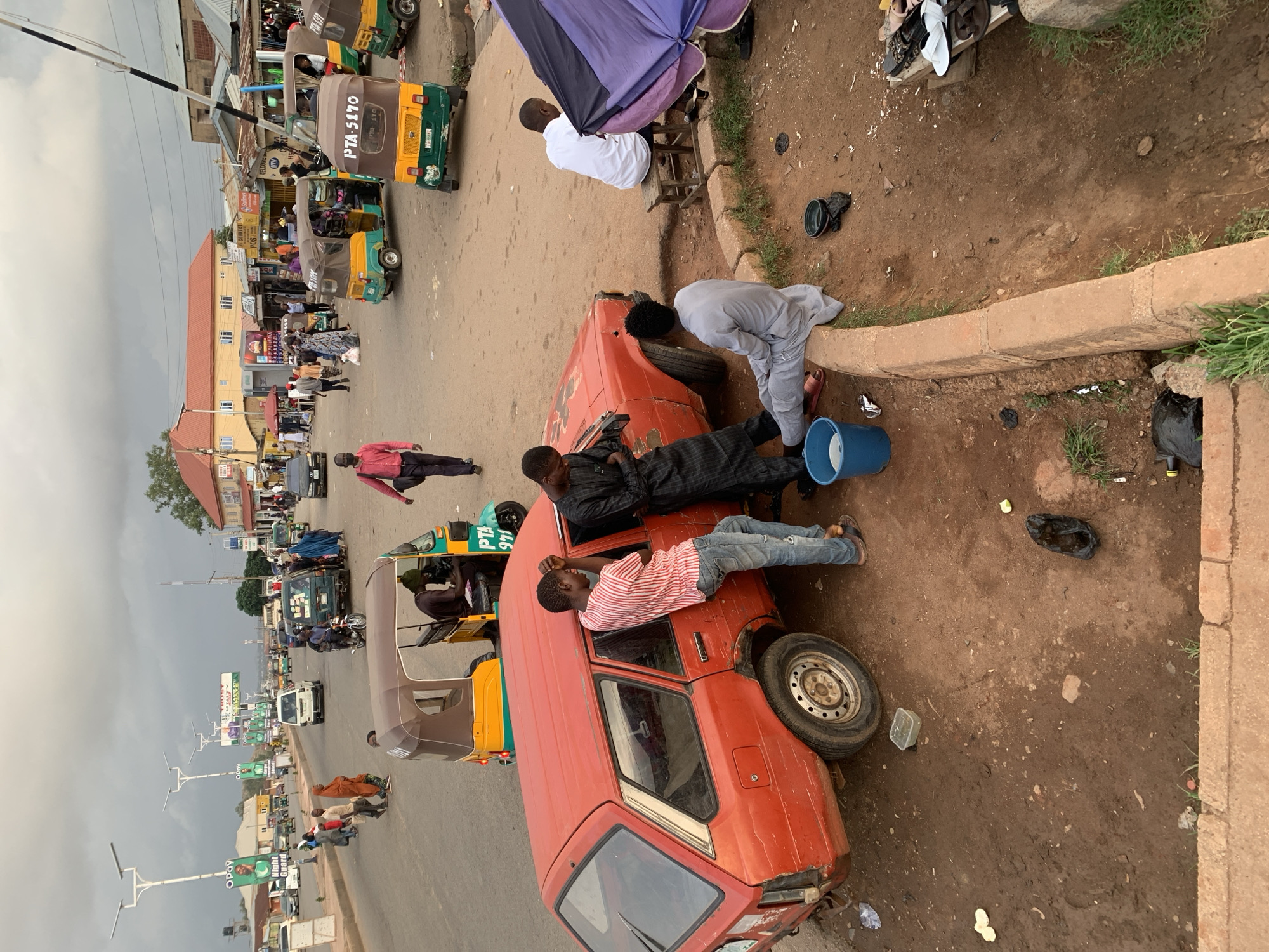 In Gura Top, Nigeria, an orange car is parked on the side of a road, a blue bucket beside it, near a sidewalk curb. Three men are situated near the car, one leaning over the car, another standing near the open driver side's door and washing, and another lying on the hood. A fourth man sits on a stool under a purple umbrella near a cart. The road extends into the distance, showing cars, tricycles, and pedestrians. Street signs and vendors are visible along the road. The sky is overcast. The ground is dirt.