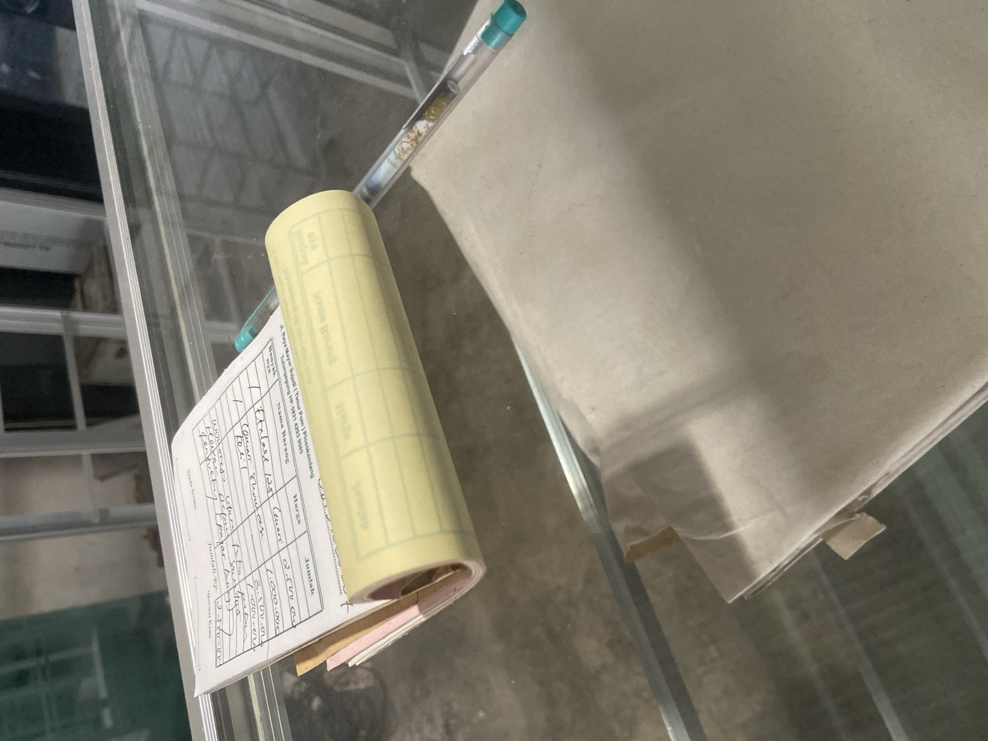 The image shows a close-up view of a receipt with handwritten entries, a pen, and what appears to be a stack of papers on a glass surface, possibly a display case. The receipt has columns for "Nama Barang" (Item Name), "Jumlah" (Quantity), "Harga" (Price), and "Jumlah Rp." (Total Rupiah). Handwritten items include "Akwel Last Cuam" with a quantity of 1 and a price of 2.500.000, "Baterai Sharpen" with a quantity of 1 and a price of 1.000.000, "Elang Menelan" with a quantity of 1 and a price of 500.000, and "Obat-obatan" with a quantity of 3 and a price of 150.000. The receipt also has "Hormat Kami" (Sincerely) written at the top. A blue and green pen is resting on the receipt, extending towards a rolled-up yellow form that seems to be a carbon copy or another type of receipt roll. The background is blurred but suggests an indoor environment with metal shelving or framing. The text at the bottom of the receipt indicates the location: "Jl. Raya Metro Sukadamai Plosokandang, Tulungagung, Hp. 0821 4253 9968." This strongly suggests the location is Plosokandang, Tulungagung, Indonesia, likely within a retail or business establishment.