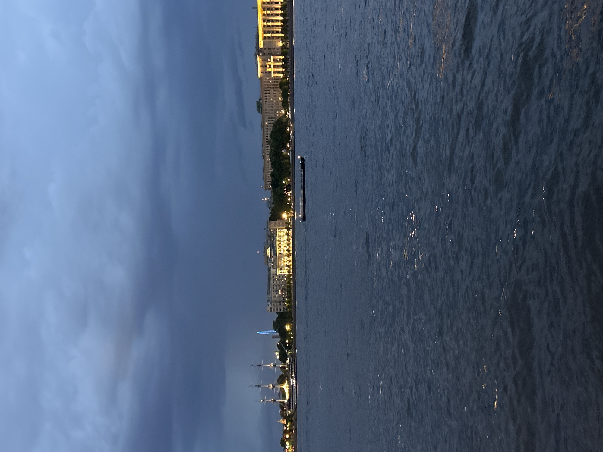 The image displays an urban waterfront scene in St. Petersburg, Russia, at dusk or night. The sky is dark blue-grey and heavily overcast with visible cloud formations. In the foreground and midground, a wide body of dark blue water, likely the Neva River, shows rippling surfaces with sparse reflections of distant lights. Along the distant left bank, a large, multi-masted sailing ship, illuminated with warm white lights, is present. Behind this vessel and further in the distance, the prominent, slender, conical structure of the Lakhta Center skyscraper is distinctly visible, illuminated with a bright blue light. The right bank features a continuous line of large, classical-style buildings, brightly lit with warm yellow-orange light that highlights their architectural details. A strip of dark trees is discernible along the embankment in front of these buildings. A dark, flat-decked cargo vessel or barge is situated in the mid-ground of the water, moving horizontally across the frame. No individuals are explicitly discernible.