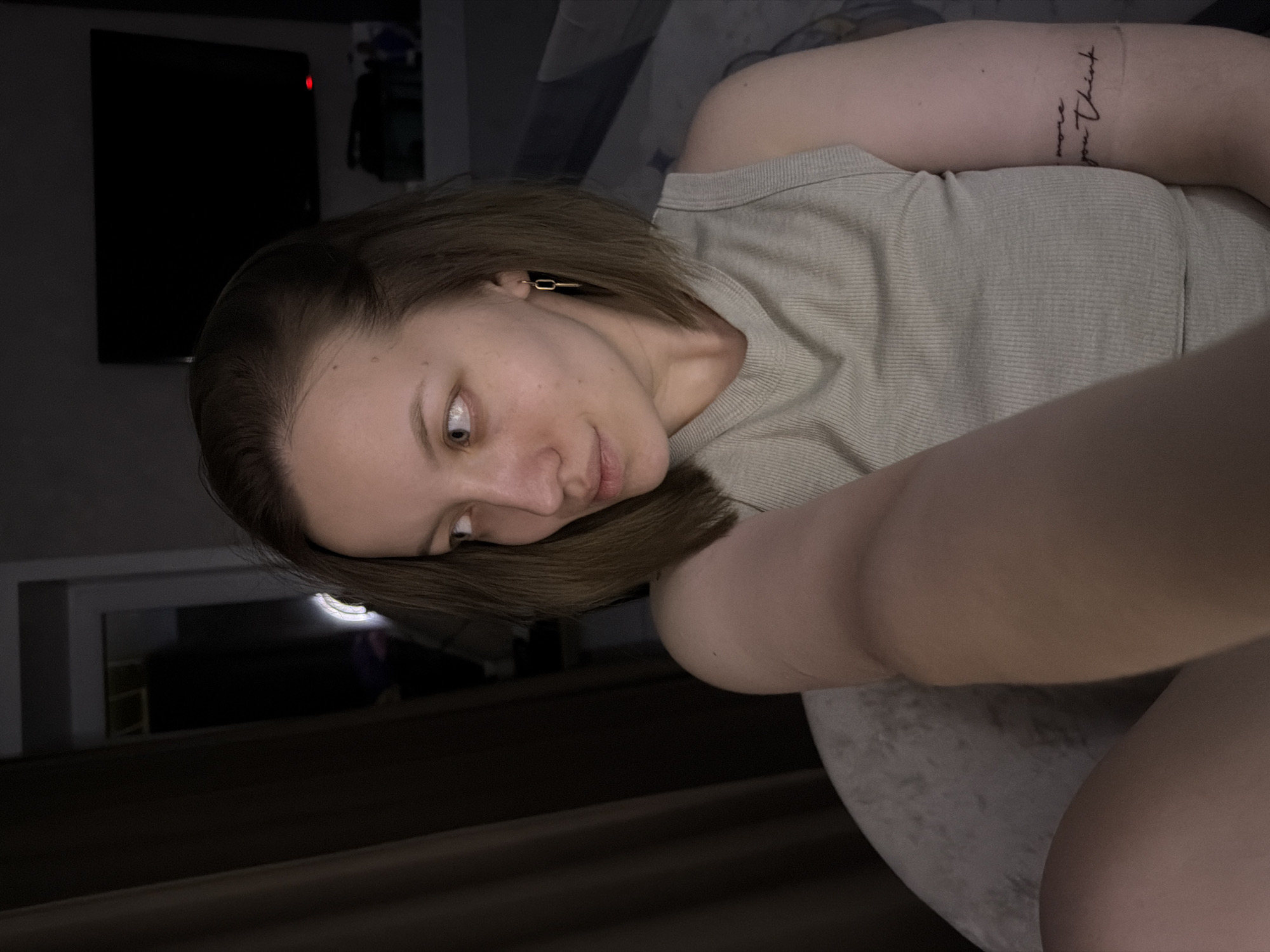 A young woman with light brown hair and fair skin is shown taking a selfie indoors in Reshetnikovo, Russia. She is wearing a light-colored, ribbed tank top and has a gold-colored rectangular link earring in her left ear. Her head is tilted, and she looks towards the camera with a neutral to slightly pensive expression. On her right forearm, a tattoo is visible, reading "more you think". The scene is dimly lit, suggesting evening or night. In the background, a dark flat-screen television with a small red indicator light is mounted on a wall. A light source reflects in her right eye and casts a glow in the lower left background, suggesting artificial illumination in the indoor setting, possibly a private residence.