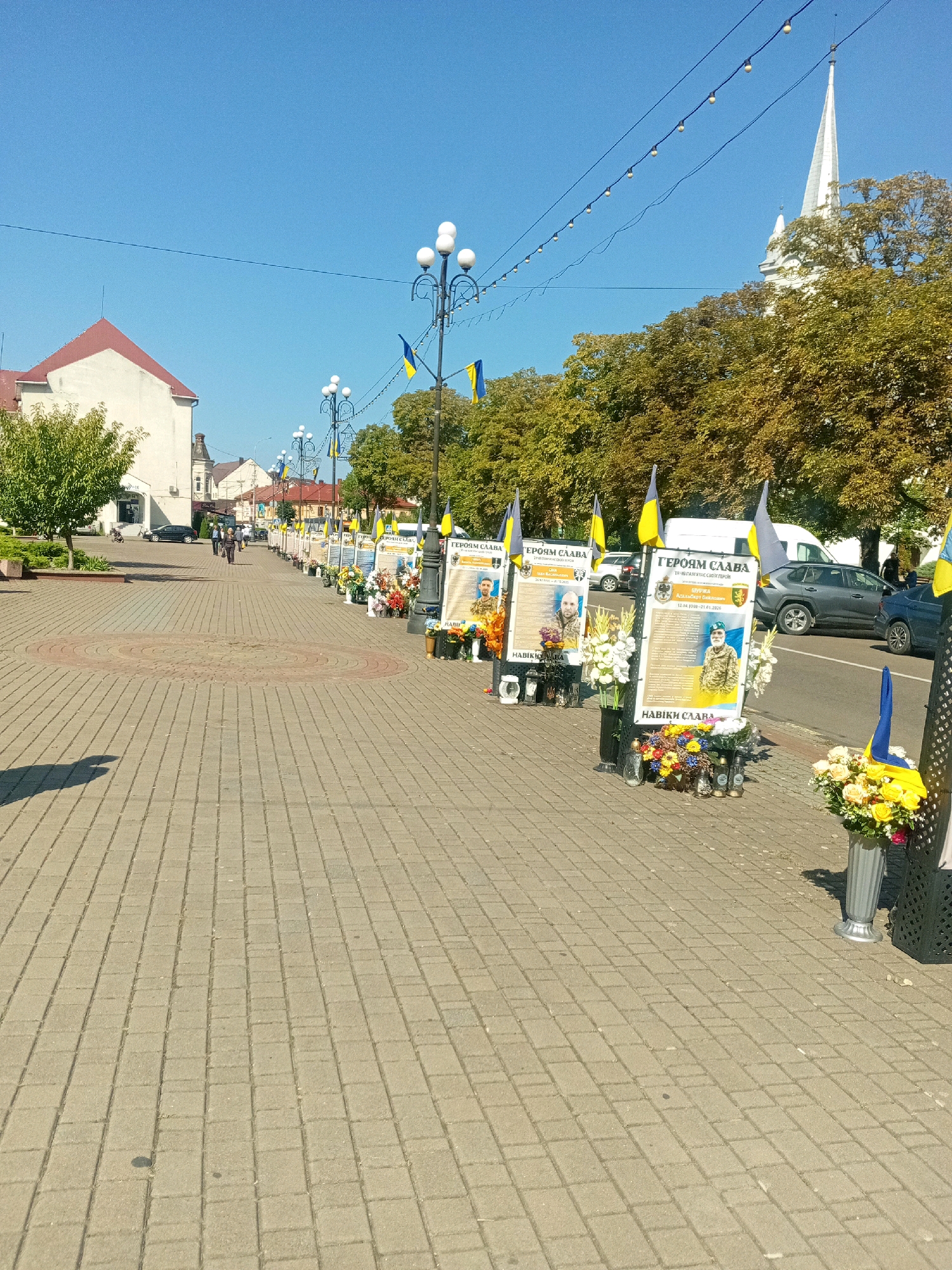 The image depicts a city street scene in Tiachiv, Ukraine, likely a memorial. Along the sidewalk, a series of framed portraits are displayed, each adorned with flowers and candles, suggesting they are portraits of fallen individuals. Ukrainian flags are prominently featured, some attached to the display stands and others held aloft. The street is paved with cobblestones, and several lamp posts with decorative lights are visible. Buildings, including a white building with a red roof and a white church steeple, line the street. Trees with green and yellow foliage are interspersed with the buildings. Several cars are parked along the street, and a few people are walking in the distance. The sky is clear and blue.