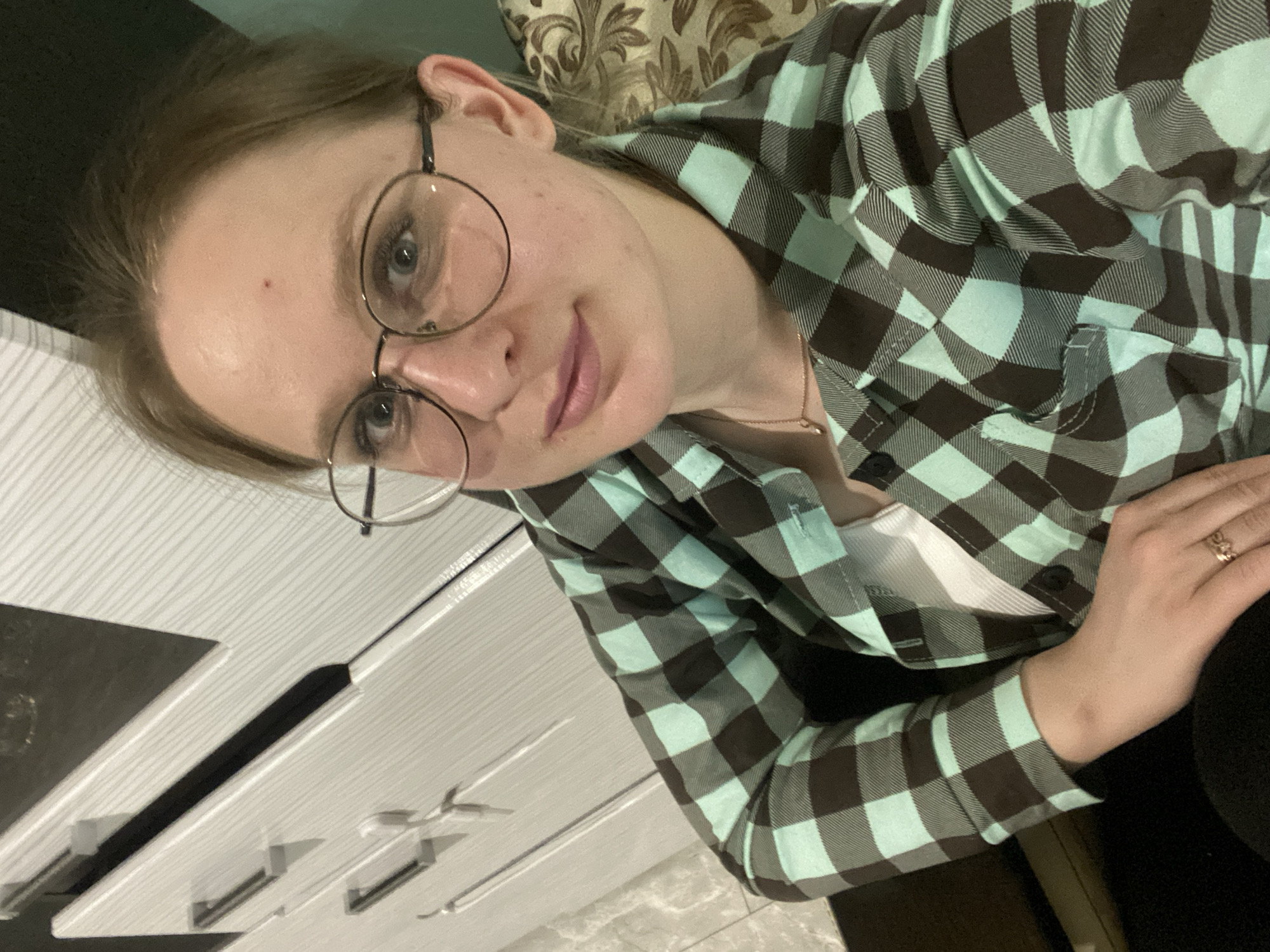 A medium shot shows a woman wearing round eyeglasses and a brown and light blue plaid shirt with a white undershirt. She is indoors, and part of a white dresser is visible on the left. She is wearing a gold ring on her right hand. A gold necklace is around her neck. The location is unspecified.