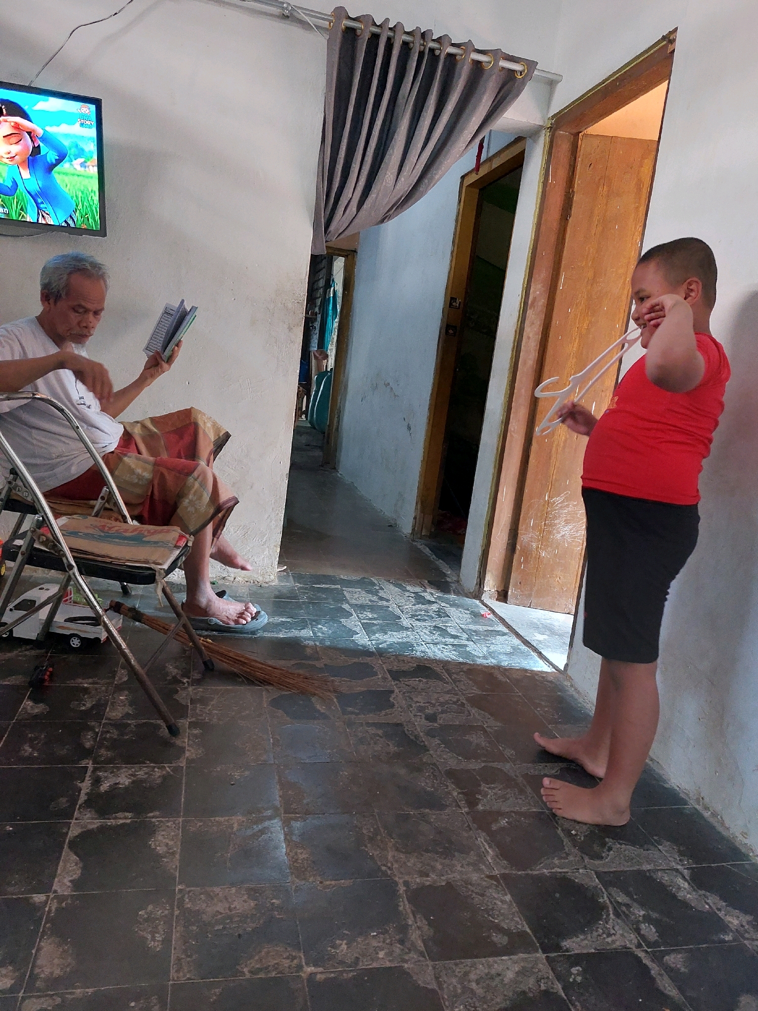 In Sarangan, Indonesia, a young boy in a red t-shirt and black shorts stands near a wooden door, holding a white hanger and touching his face. To the left, an elderly man with gray hair sits in a folding chair, reading a book and wearing a white t-shirt and a patterned sarong. A television screen displays a cartoon. The floor is tiled with dark, worn tiles.