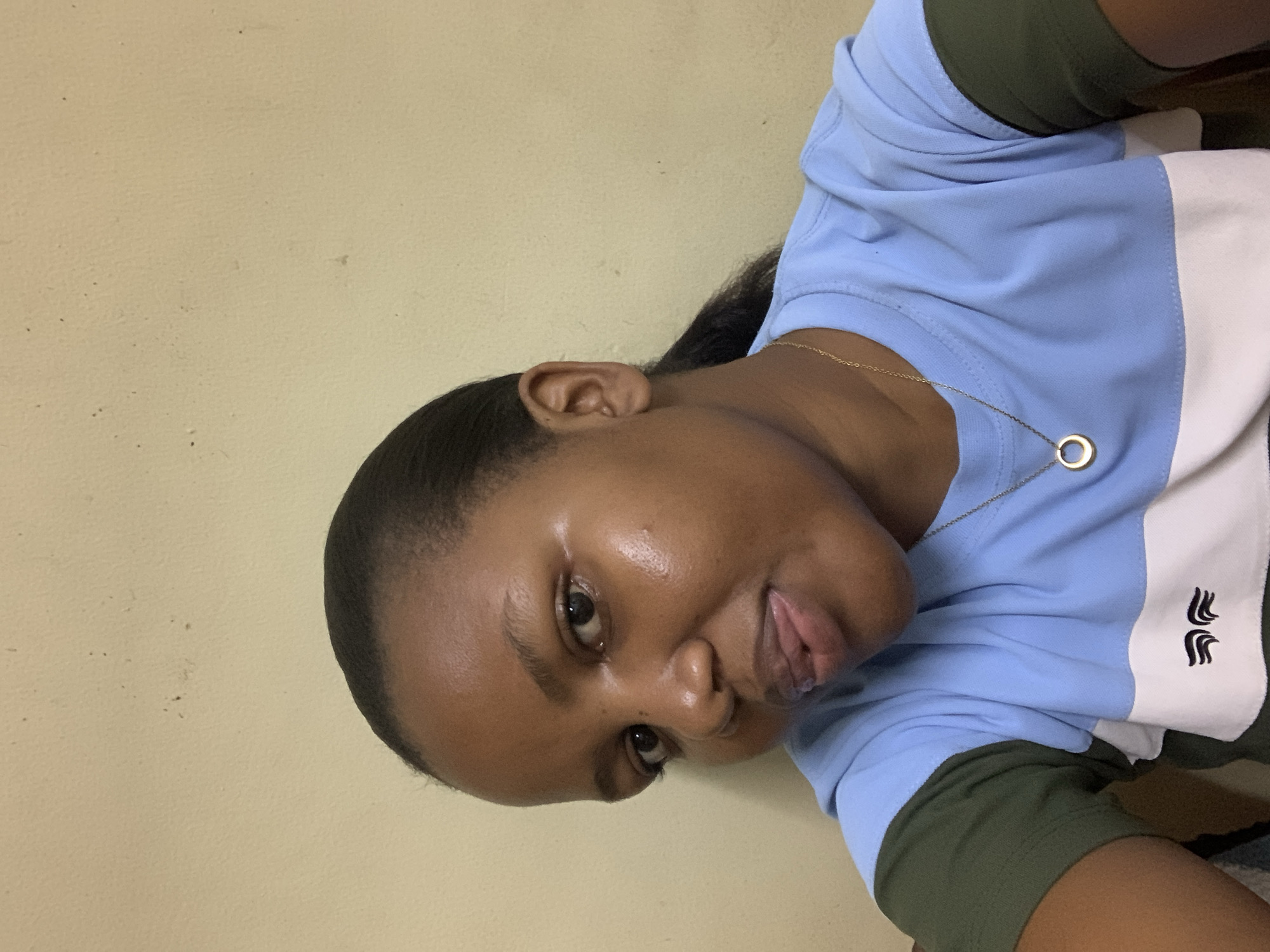 A close-up, angled portrait features a young person, likely female, against a plain, light-colored wall, taken indoors in Dar es-Salaam, Tanzania. The subject's head is tilted, and they gaze directly at the camera with a relaxed, slightly playful expression, their tongue visible. Their dark hair is pulled back from their face, and their ear is visible.
The person wears a collared shirt with distinct color blocking: light blue across the shoulders and upper body, a central white stripe, and an olive-green section below. On the white stripe, a dark embroidered logo depicts two stylized, intertwined shapes resembling flames or feathers. A slender, gold-colored chain necklace with a small, circular pendant adorns their neck. The background wall shows minor textured imperfections. The lighting appears even, suggesting an indoor setting.