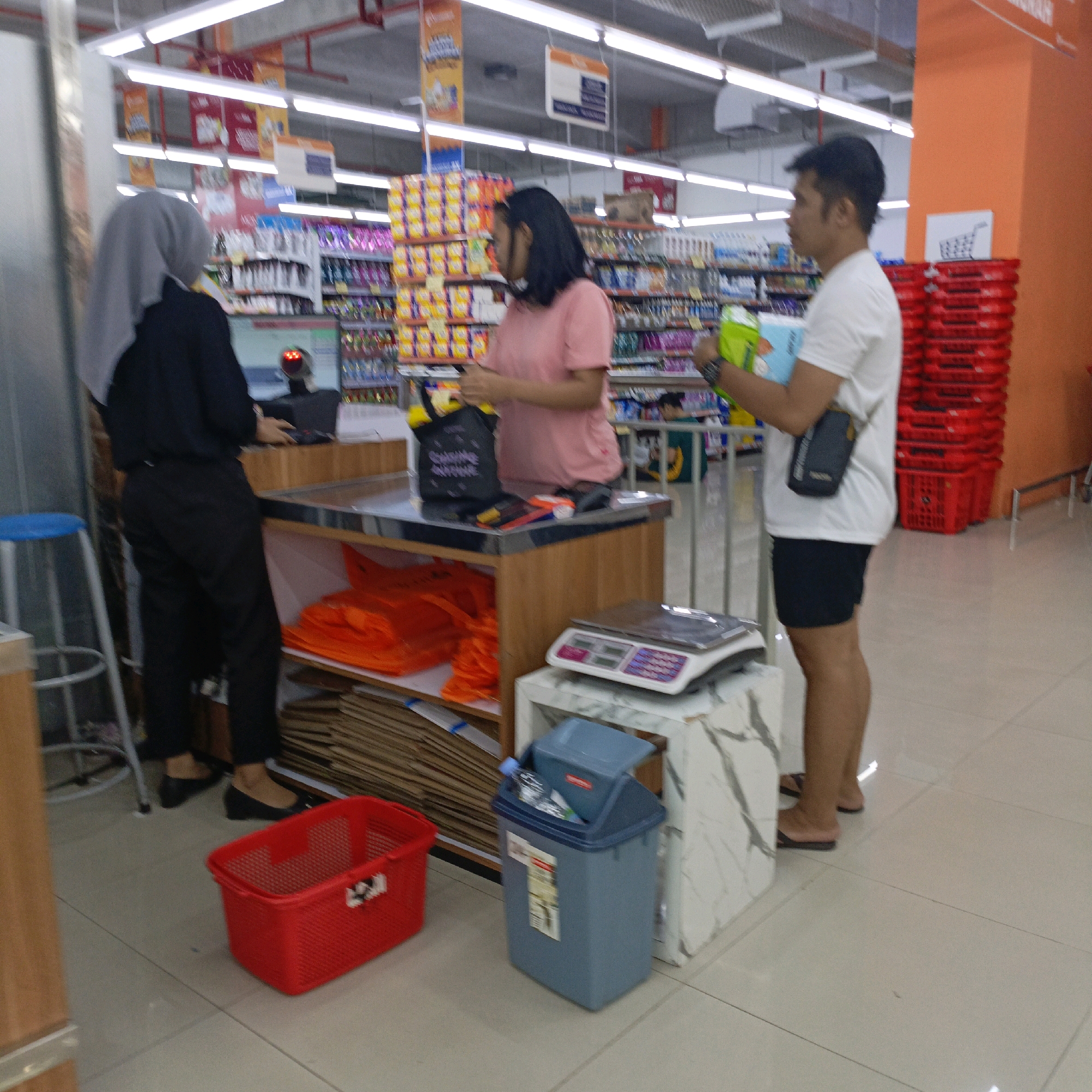 This image depicts a checkout counter area within a supermarket in Salatiga, Indonesia. Two individuals are visible: a woman wearing a grey hijab and black clothing, operating a cash register, and a man in a white t-shirt and dark shorts holding a green and blue object. To the left of the woman is a blue stool. In front of the counter, a red plastic basket sits on the floor, next to a grey trash bin. Behind the counter, red shopping baskets are stacked on an orange wall. The environment suggests a commercial retail space with shelves stocked with products in the background.