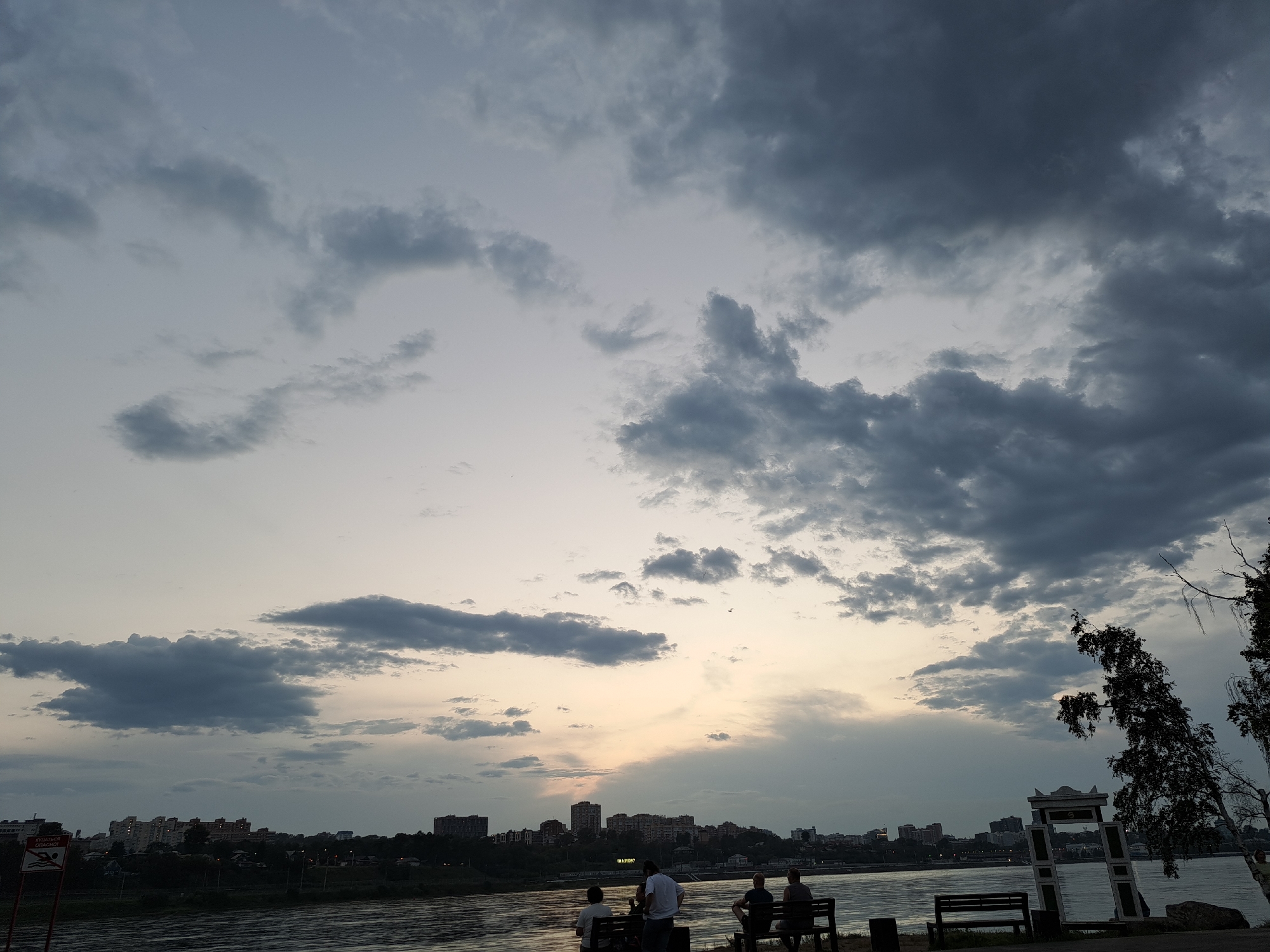 The image depicts a waterfront scene in Irkutsk, Russia, with a city skyline in the distance. The sky is filled with dramatic, dark clouds, suggesting an approaching storm or the aftermath of one. In the foreground, several people are seated on benches along the embankment, observing the river and the city. The river itself appears calm, reflecting the muted light of the overcast sky. The city skyline features a mix of residential buildings and some taller structures, illuminated by lights that suggest it is either dusk or dawn. To the right of the image, a decorative archway stands near some trees, adding an architectural element to the natural landscape. A warning sign in Russian, with symbols indicating danger, is visible on the left side of the frame, suggesting this area might be designated for swimming or water activities but has potential hazards. The overall atmosphere is serene yet somewhat somber due to the heavy cloud cover.