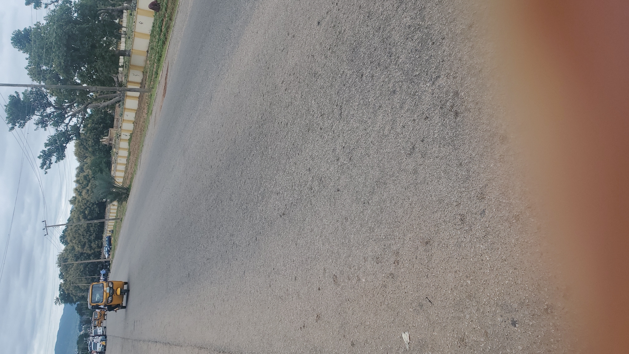 An outdoor, eye-level, medium shot captures a street scene in Jalingo, Nigeria, under a cloudy sky. In the foreground, the asphalt road stretches into the distance, with a blurry, light brown area at the bottom. To the left, a yellow auto-rickshaw, or Keke Napep, is in motion, with a person visible in the driver's seat and a passenger behind them. Several other vehicles, including cars and more Keke Napeps, are visible on the road further ahead. Along the right side of the road, a low, yellow-and-white striped wall marks the boundary of a property lined with trees. Some of these trees are in bloom with light-colored flowers, while others are leafy and green. A utility pole with wires stands near the left side of the road, and another is visible further back. In the background, a mountain range is faintly visible. A person can be seen near the wall on the right side of the road.