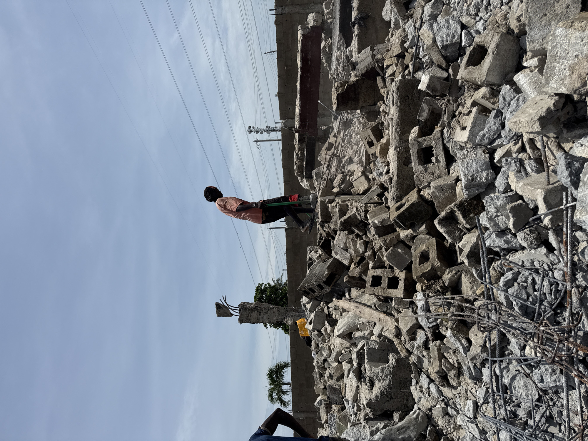 A man stands atop a pile of rubble in Ado, Nigeria. The rubble consists of broken concrete blocks and exposed rebar. The man is wearing a peach-colored shirt, dark pants, and a black head covering. He appears to be operating some kind of machine with a green handle. The background features a partly cloudy sky, utility poles, and a partially demolished building. A partial view of another person's lower body is visible at the bottom of the image.