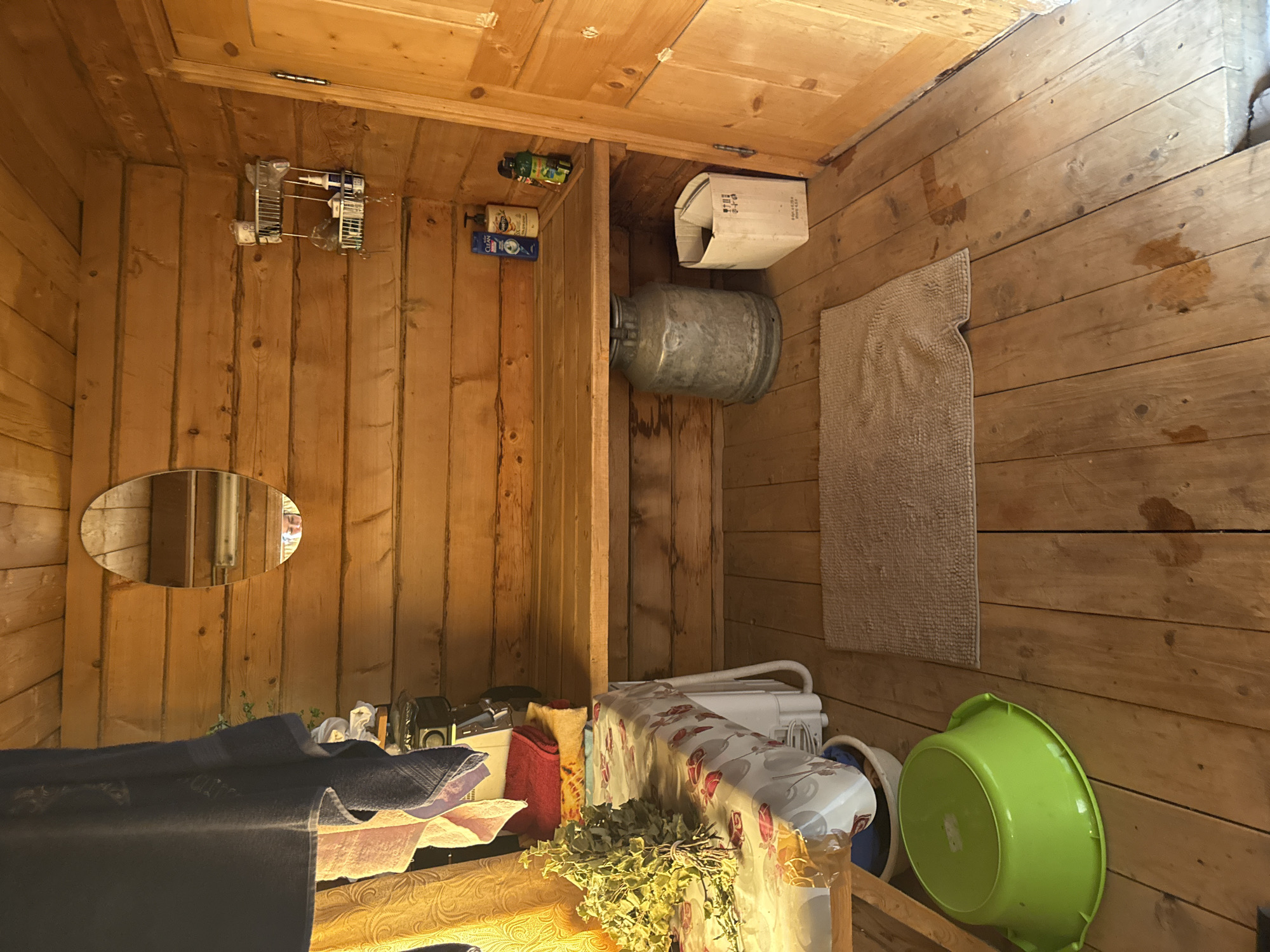 The image depicts the interior of a wooden room, likely a Russian banya or sauna, located in Чуй-Атасево, Russia. The walls and ceiling are constructed from horizontal wooden logs. A wooden bench runs along the back wall, above which is a narrow shelf holding several bottles of toiletries and a small wire rack with additional items. A large oval mirror is mounted on the log wall above the shelf.
To the left of the bench, a table is covered with a patterned tablecloth. On the table sits a bunch of dried leafy branches, possibly for use in a sauna. Behind the table, several folded towels and what appears to be a small appliance are visible. A yellow curtain hangs to the left of the table.
On the wooden floor, a light brown, rectangular mat lies in the center of the room. To the left of the mat, a large, bright green plastic basin is placed on the floor. Beneath the bench, a large, galvanized metal milk can stands next to a white cardboard box. The wooden floor shows some water stains and appears worn. A wooden door with a simple design is visible on the right side of the image. The overall impression is of a functional, rustic bathing space.