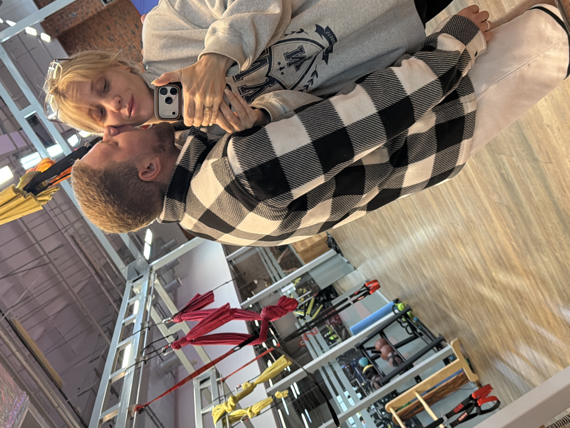 This media file captures an intimate moment between a man and a woman in what appears to be an indoor fitness studio or gym in Chaikovsky, Russia. The image is oriented with the subjects leaning towards each other from what would be the left and right sides if correctly rotated.
The woman, with blonde hair and glasses pushed onto her head, wears a light grey hooded sweatshirt adorned with a dark, stylized crest featuring letters that resemble "M" and "V." She holds a dark-colored smartphone in a clear and orange case, seemingly taking a selfie. A ring is visible on her left ring finger. Her eyes are closed or looking down, and she exhibits a gentle, affectionate expression.
The man, with light brown hair and a short beard, wears a black and white buffalo plaid flannel shirt. He leans in towards the woman, their faces nearly touching, suggesting a kiss or close embrace. Their hands are also close, possibly intertwined, emphasizing their connection.
The background reveals an industrial-style interior. The ceiling features exposed metal beams, ducts, and fluorescent lighting. Below, various gym equipment is visible, including multiple sets of red and yellow aerial silks hanging from a metal frame structure. Further back, storage racks hold items like medicine balls, foam rollers, and suspension trainers with red handles, though specific text on these items is largely illegible. The floor is made of light-colored wood or laminate, and a reflective surface creates distorted reflections of the equipment on the right side of the frame. The overall atmosphere conveys a moment of warmth and affection within a functional setting.