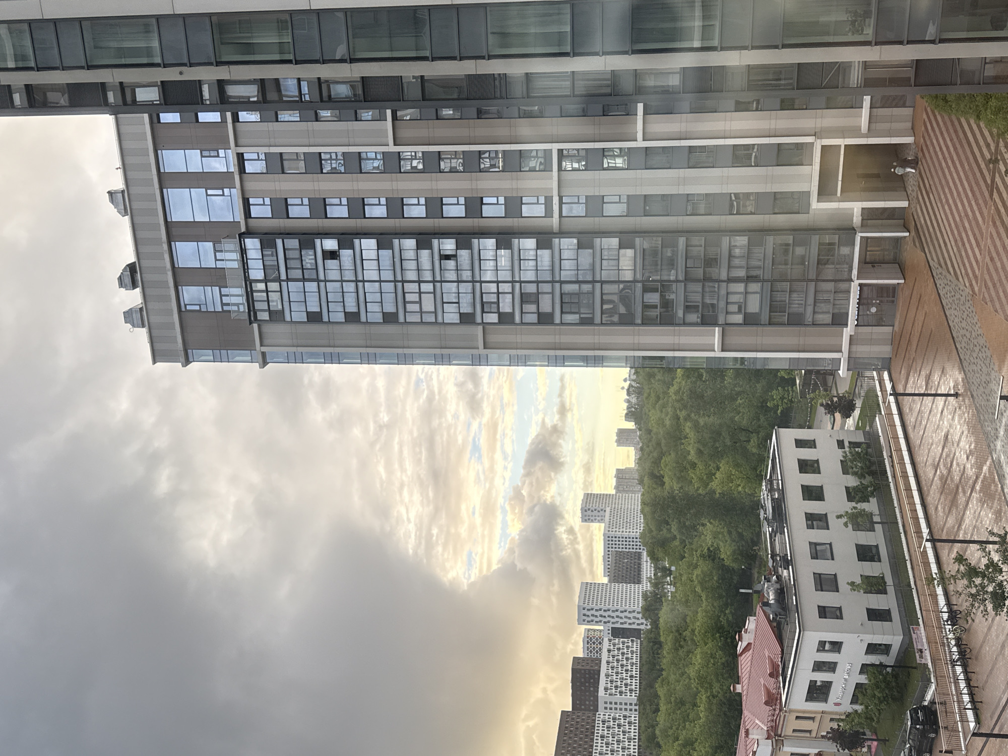 The image shows a mixed urban and natural landscape in Khimki, Russia. A tall, modern building with reflective windows dominates the view, juxtaposed against a cloudy sky. In the distance, several other high-rise buildings are visible, interspersed with green trees suggesting a park or forested area. In the foreground is a view of the street and a building with fewer floors. A sidewalk runs beside this building, with some bike racks. The overall scene has a slightly overcast, possibly post-rain ambiance.