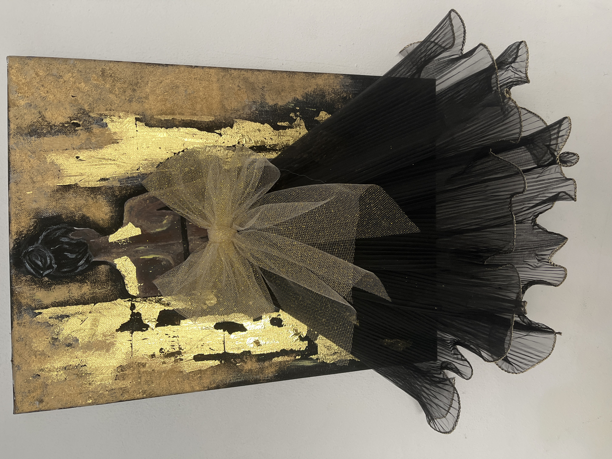 The image depicts a piece of art, likely a mixed-media canvas, featuring a stylized silhouette of a person from behind. The background is textured with prominent gold leafing applied in vertical, abstract strokes against a darker, possibly black, base. A large, golden tulle bow is positioned at the waist of the silhouette. Below the bow, a voluminous black skirt, constructed from pleated sheer fabric with gold trim along the ruffled edges, cascades downwards. The silhouette itself appears to be painted, with dark hair styled in an updo. The artwork is mounted against a plain white wall, suggesting an indoor setting. There is no indication of the city of Topoli, Bulgaria, or any specific location within it in this image.