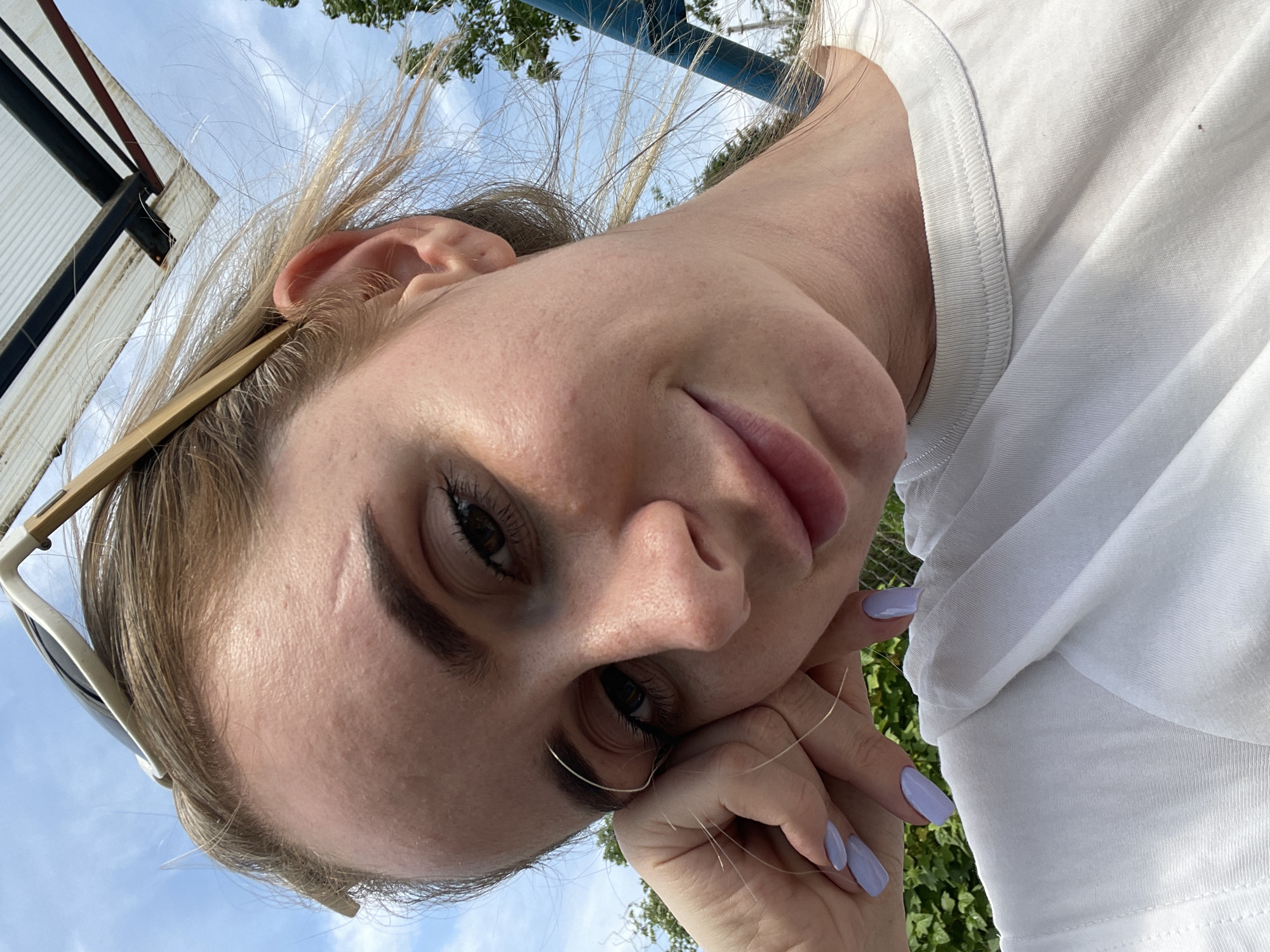 A woman is posing outdoors on a sunny day. She wears a white T-shirt and has blond hair. Her nails are painted a light purple color. She is looking directly at the camera with her head tilted. Sunglasses are perched on top of her head. The background includes a blue sky with some clouds and trees. The location is unspecified.