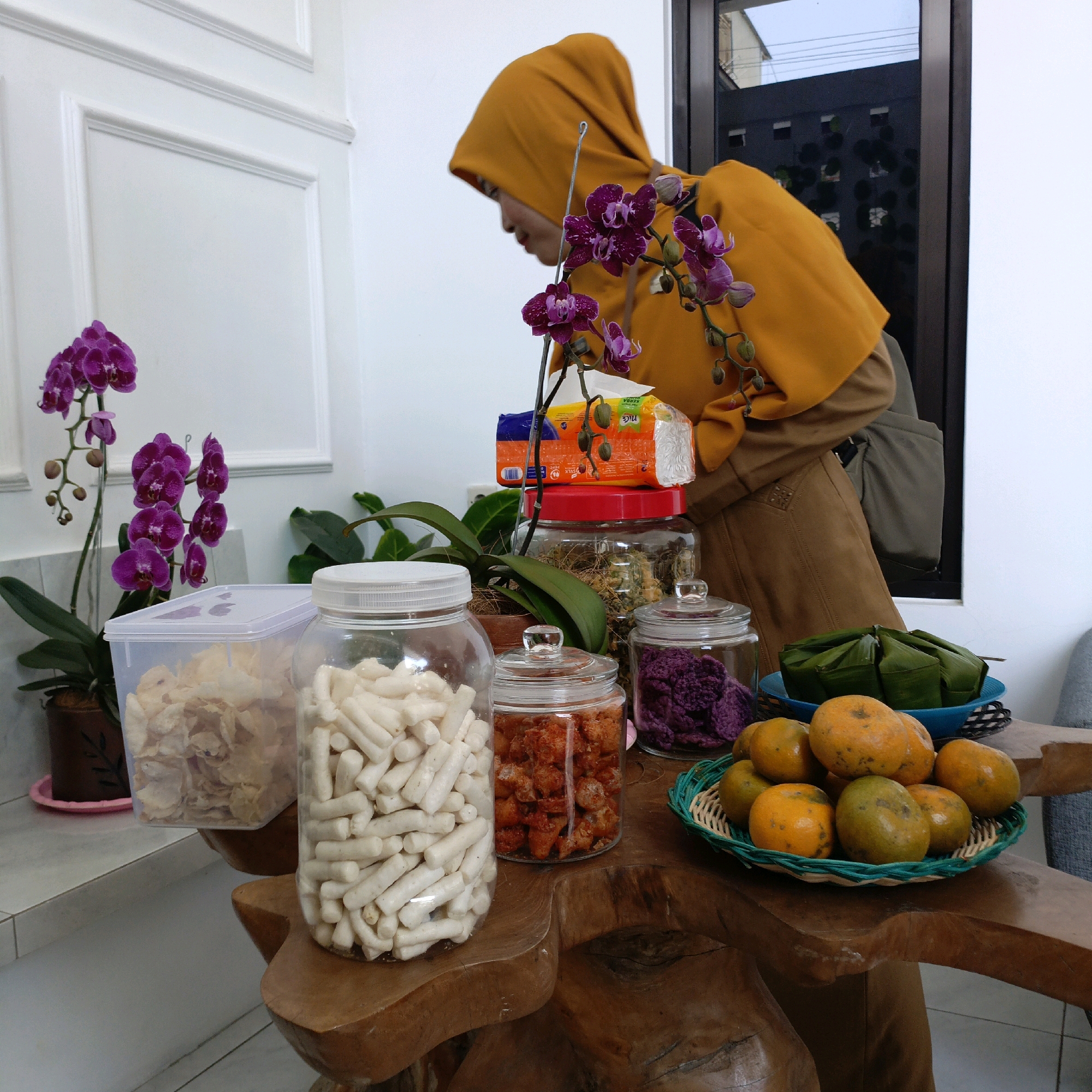 In Salatiga, Indonesia, a woman wearing a mustard-yellow hijab and brown attire is captured in an indoor scene. She is leaning forward, seemingly interacting with items on a wooden table. The table is adorned with several clear glass jars containing snacks: one with light-colored, tubular snacks; another with reddish-orange snacks; and a third with what appears to be purple shredded ingredients. A clear plastic container filled with light-brown, flaky snacks is also present. Further additions to the tabletop arrangement include a blue plate holding several green-wrapped food parcels and a woven basket brimming with oranges. Two potted orchid plants with vibrant purple flowers add a touch of natural beauty to the composition. In the background, a white wall with decorative molding and a dark-framed window offer glimpses of the surrounding environment, including hints of an outdoor space.