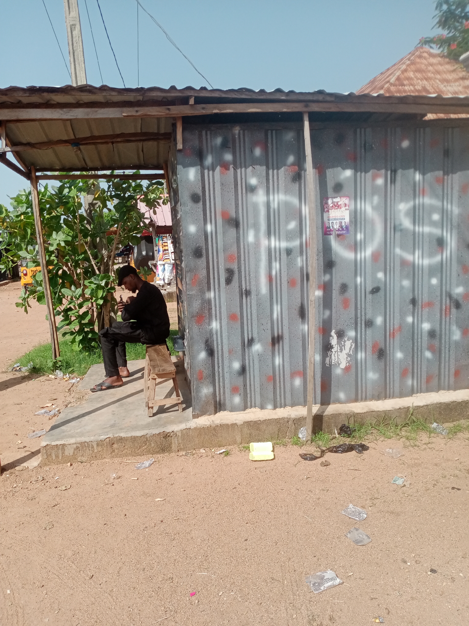 In Jalingo, Nigeria, a young man wearing a black shirt, black pants, and a black cap sits on a wooden stool under a covered, open-air structure. He is looking down at his phone, which he holds in his right hand. The structure has a corrugated metal roof and walls made of corrugated metal panels painted with a pattern of gray, black, and red dots, with white spray-painted graffiti. To the left of the man, a green tree with lush foliage grows, and in the background, a yellow trishaw is partially visible. The ground in front of the structure is dirt and debris, with scattered pieces of trash including plastic bags and wrappers. A concrete platform runs along the base of the structure. The sky above is clear and blue.