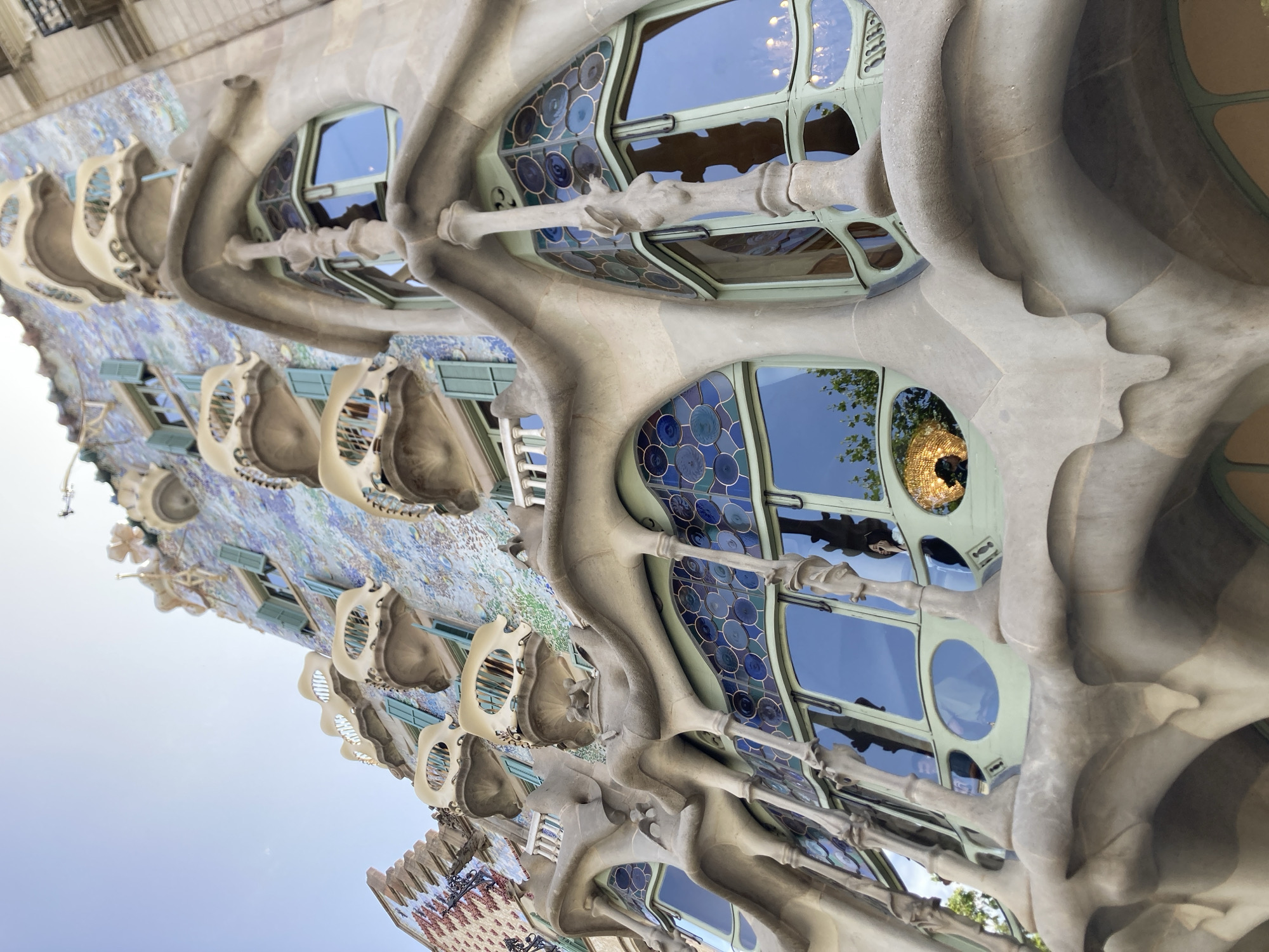 The image shows a close-up view of the facade of Casa Batlló in Barcelona, Spain. The architecture is organic and flowing, with curved lines and bulbous shapes reminiscent of natural forms. The facade is adorned with colorful mosaic tiles and decorative elements that resemble bones and skulls. The windows are uniquely shaped and feature stained glass with circular patterns in shades of blue and green. Sunlight reflects off the windows, creating bright highlights and casting shadows that emphasize the three-dimensional quality of the structure. The sky is visible in the background, suggesting an outdoor setting during the daytime. The image is oriented vertically, giving the impression of looking up at the building. The building's distinctive style is characteristic of Antoni Gaudí's modernist architecture.