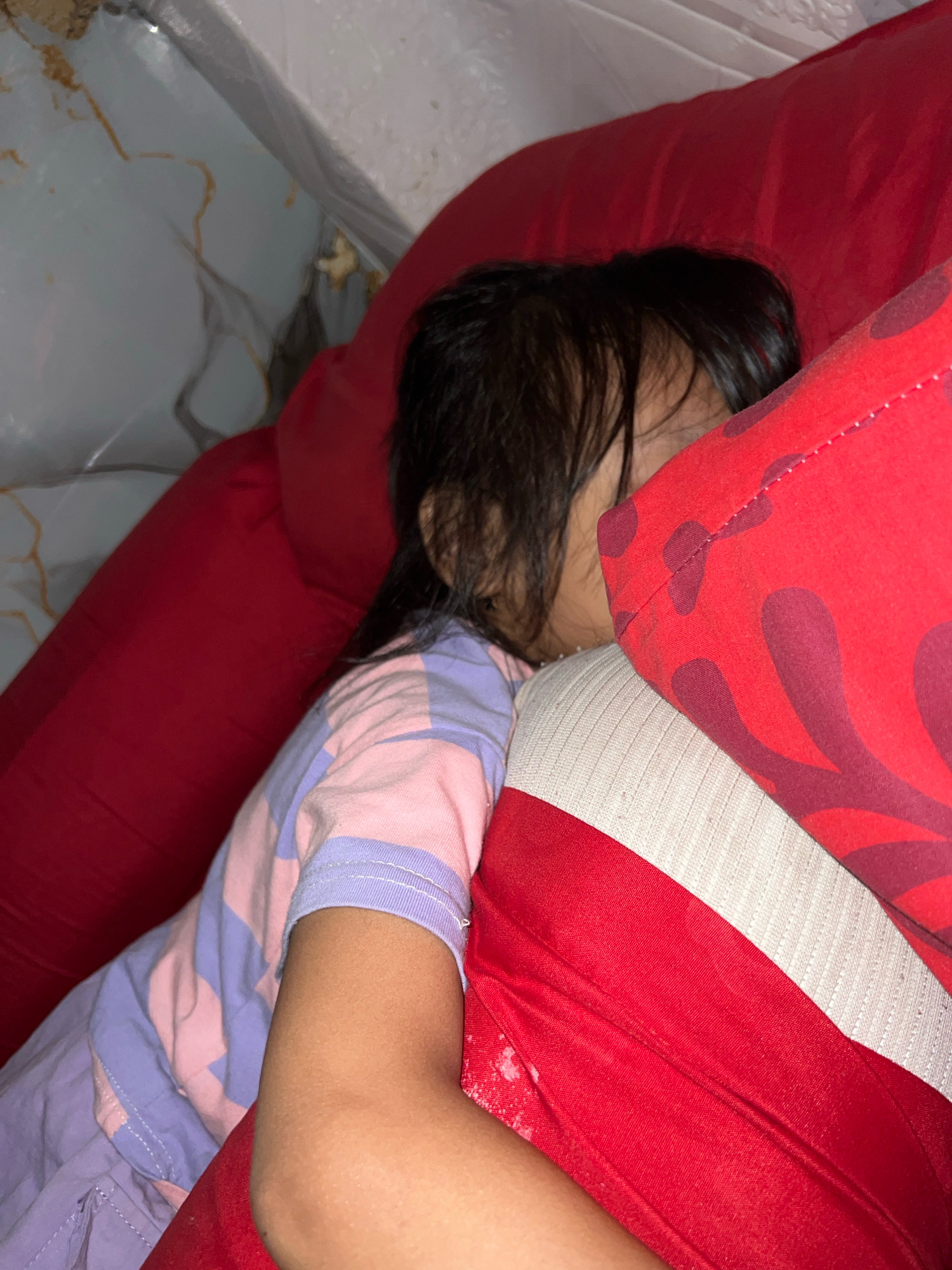 A young child is sleeping on a red couch in a home in Kecamatan Pati, Indonesia. The child is lying on their side, with their face partially obscured by their hair and a red pillow. They are wearing a pink and purple striped t-shirt. The couch appears to be made of a soft fabric, and there are other red pillows visible in the background. The room is dimly lit, and the overall impression is one of comfort and relaxation.