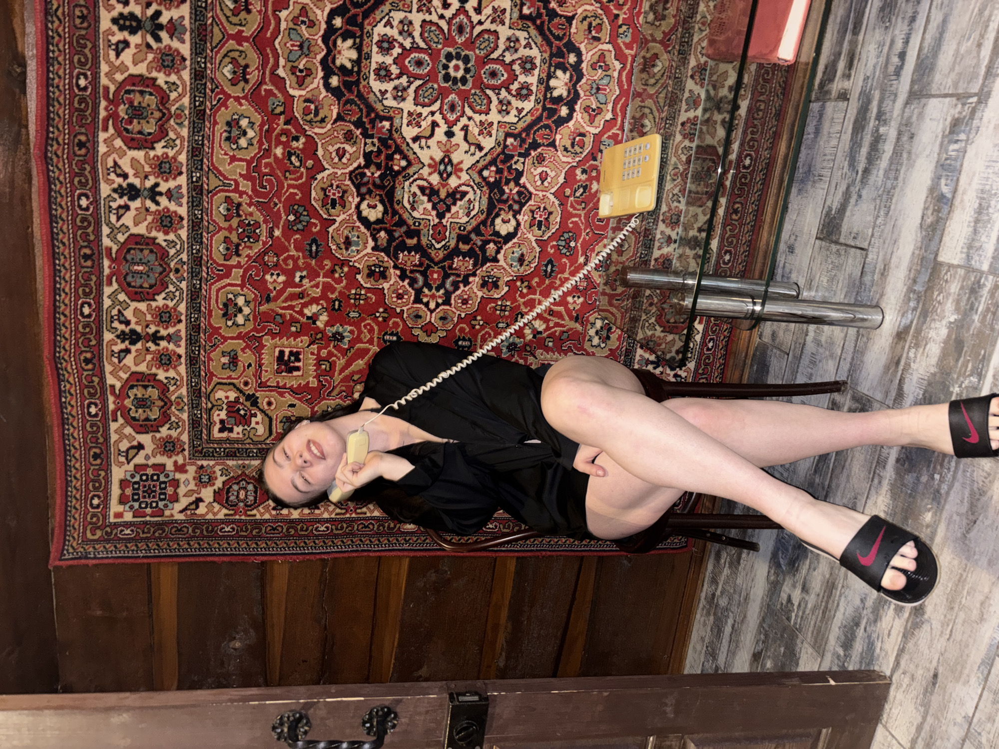 A young woman in a black robe and black Nike slides is lying on a red patterned rug in a room with a glass-topped table and wood-paneled walls. She is holding a yellow landline telephone receiver to her ear. The floor is a light wood. The location is Perm, Russia.