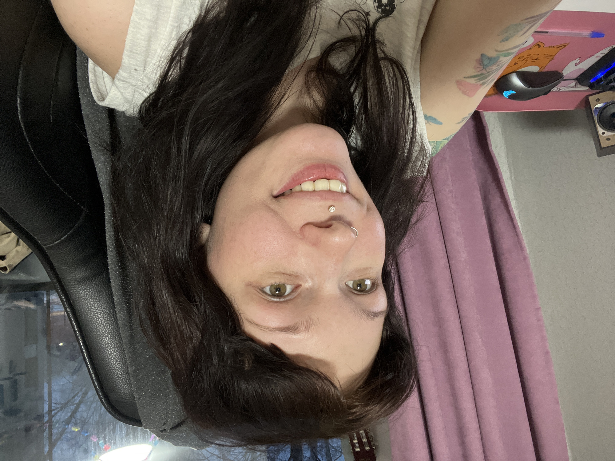 A woman with dark hair is looking at the camera. She is wearing a light-colored shirt and is sitting in a black chair. She has piercings on her nose and lip. Her left arm has several tattoos. Behind her is a pink curtain and a gray wall. A desk with a computer mouse, pen, and speaker are visible on the right side of the image. The presence of bare trees and a winter scene outside the window suggests the image may have been taken in Chaikovsky, Russia.