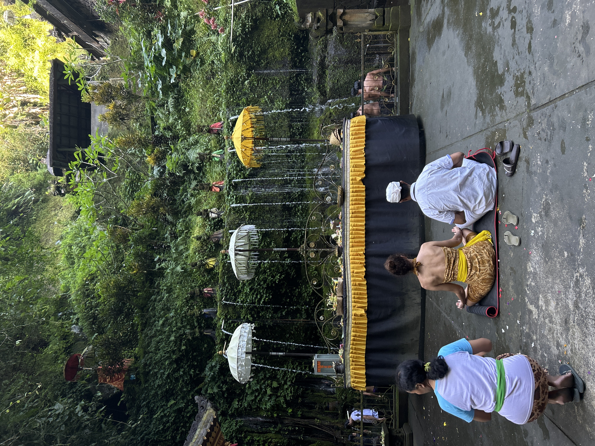 The image depicts a spiritual site in Bangli, Indonesia, likely Tirta Empul Temple or a similar water temple. Three individuals are visible in the foreground, facing away from the camera, engaged in prayer or meditation. One person, on the left, kneels with hands clasped, wearing a light blue and white shirt and a patterned skirt with a green sash. In the center, a woman in a patterned sarong and yellow sash meditates cross-legged on a mat. To her right, a man in a white shirt and white cap sits cross-legged on a mat.
Behind them is a raised altar draped in black fabric with a yellow, pleated valance. Several decorative white and yellow umbrellas are positioned above the altar, from which water streams down, creating a cascade effect. Small figures, possibly religious statues, are arranged on a ledge above the waterfalls. The background is a lush, green, densely vegetated cliff face, suggesting a natural grotto or ravine. Additional figures are visible further back, near what appears to be a bathing pool with a fence. The ground in the foreground is wet and speckled with flower petals. The overall atmosphere is serene and contemplative.