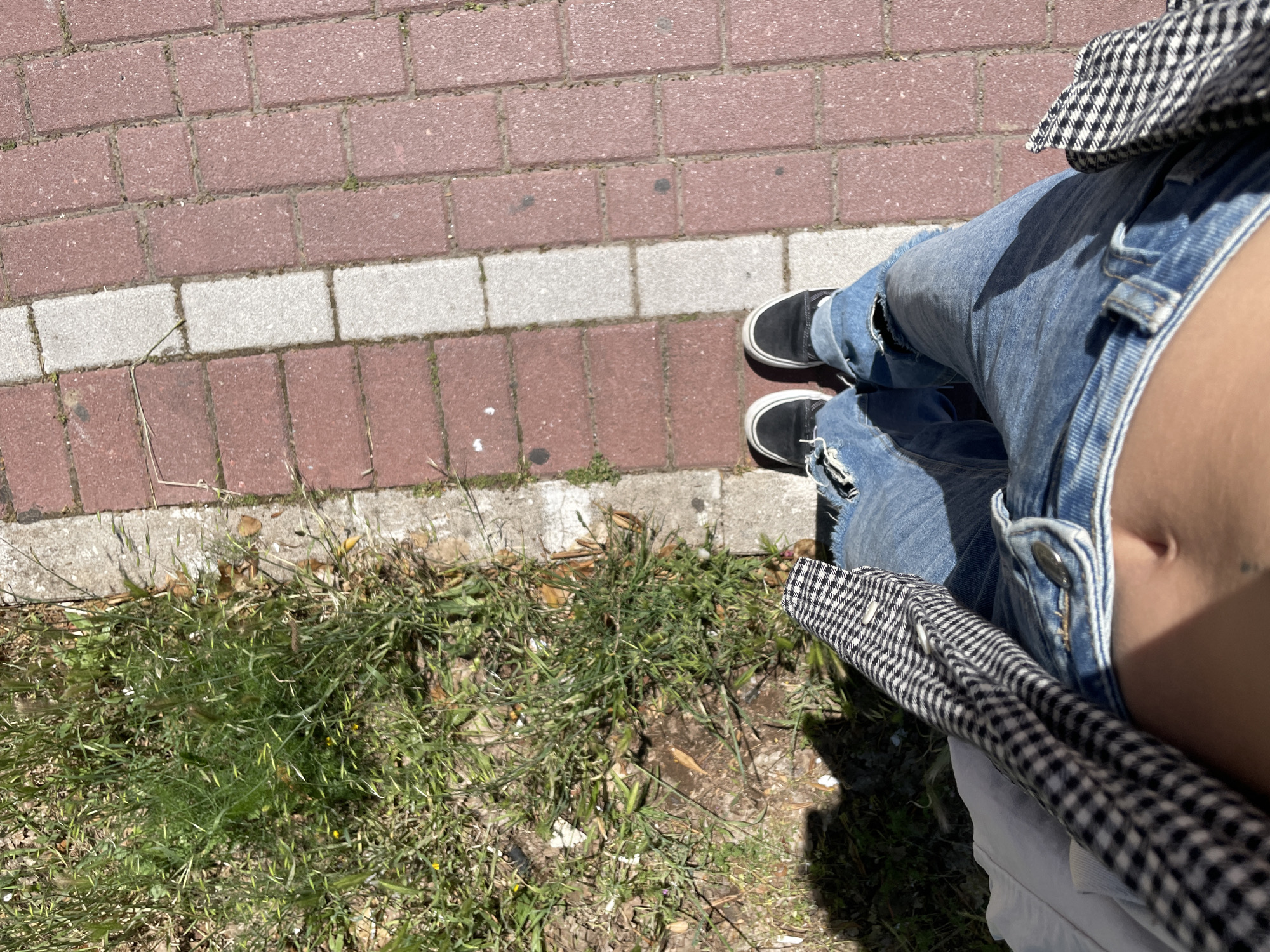 A first-person perspective photograph taken outdoors shows the lower part of a person standing on a brick walkway next to a patch of grass. The person is wearing ripped blue jeans, black slip-on shoes with white soles, and a black and white checkered garment. The walkway consists of alternating rows of red and grey bricks. The grass patch is green and uneven, with some dry leaves and dirt visible. The image is taken at a high angle, looking down. The location of the image is Rafailovići, Montenegro.