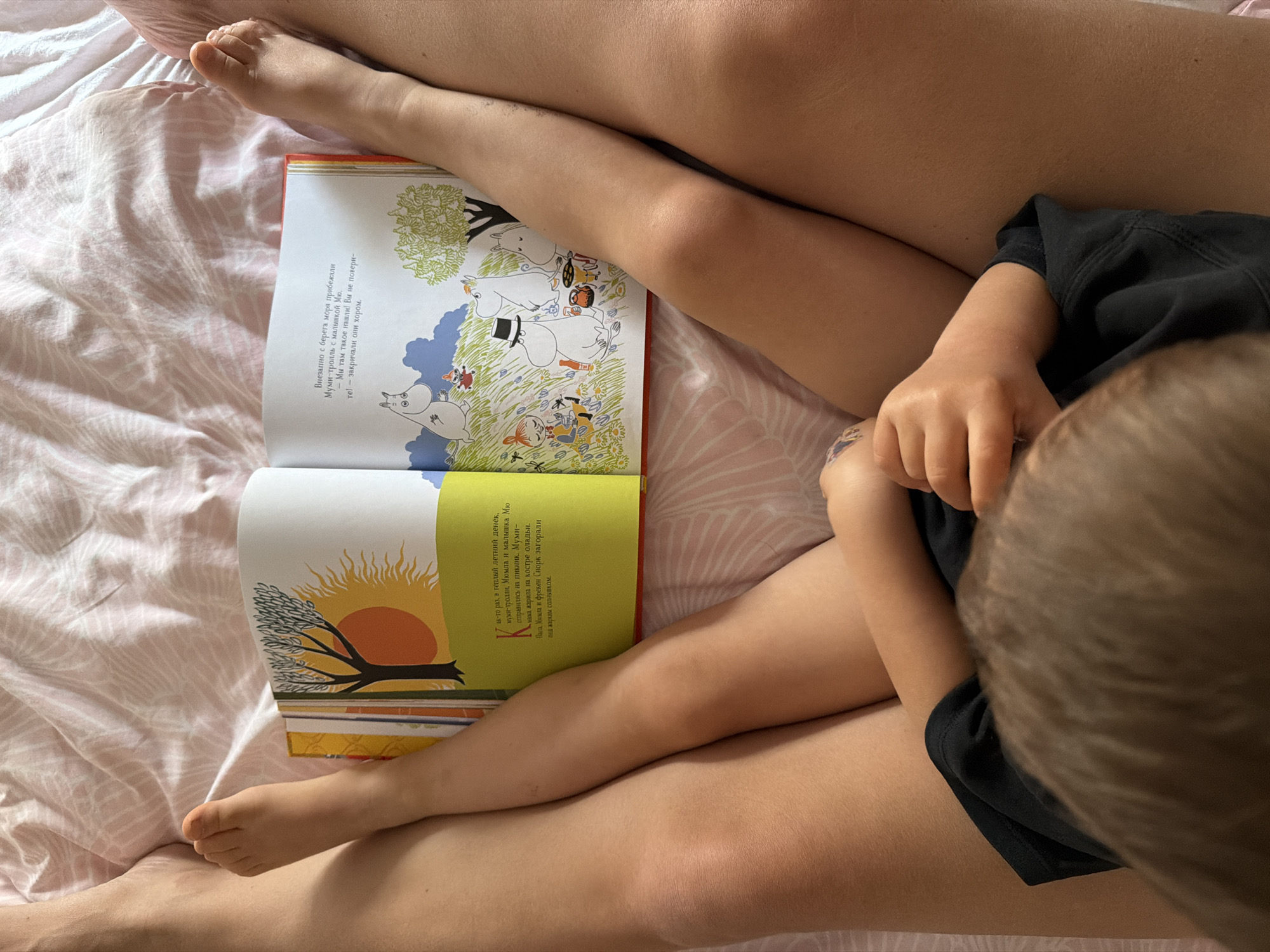 The image presents a warm, top-down view of an adult and a child sharing a cozy moment on a bed in Budva, Montenegro. Their intertwined legs and feet are prominent, with the child's smaller limbs resting between the adult's. The child's head, seen from above with light brown hair, is visible on the right, bent over an open book, suggesting a shared reading activity.
An open Moomin-themed book lies centrally, displaying colorful illustrations and Russian text. The left page depicts Moomintroll, Snorkmaiden, and other Moomin characters in a grassy landscape under a blue sky, with trees. The visible text describes Moomintroll and Little My running from the sea, excitedly exclaiming about something they found. The right page features a vibrant orange sun with rays, a tree, and more Moomin characters. Its text begins a story about Moomintrolls, Mymble, and Little My going on a picnic on a warm summer day, with Moominmamma frying pancakes and others sunbathing.
The bed is covered with a soft, light pink bedsheet featuring a subtle leaf or fern pattern. The natural lighting indicates it is daytime. The scene conveys a sense of peaceful bonding and shared storytelling.